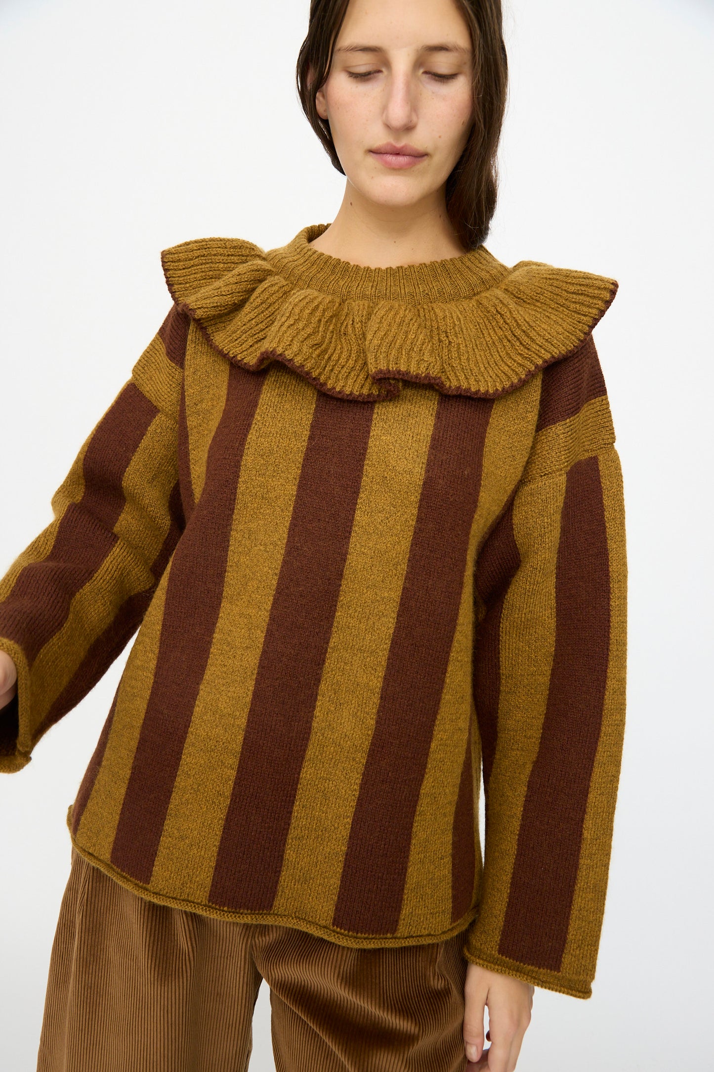 A person wearing the Cawley British Merino Wool Striped Ruffle Jumper in Goldfinch and Bordeaux with tan corduroy pants stands against a plain background.