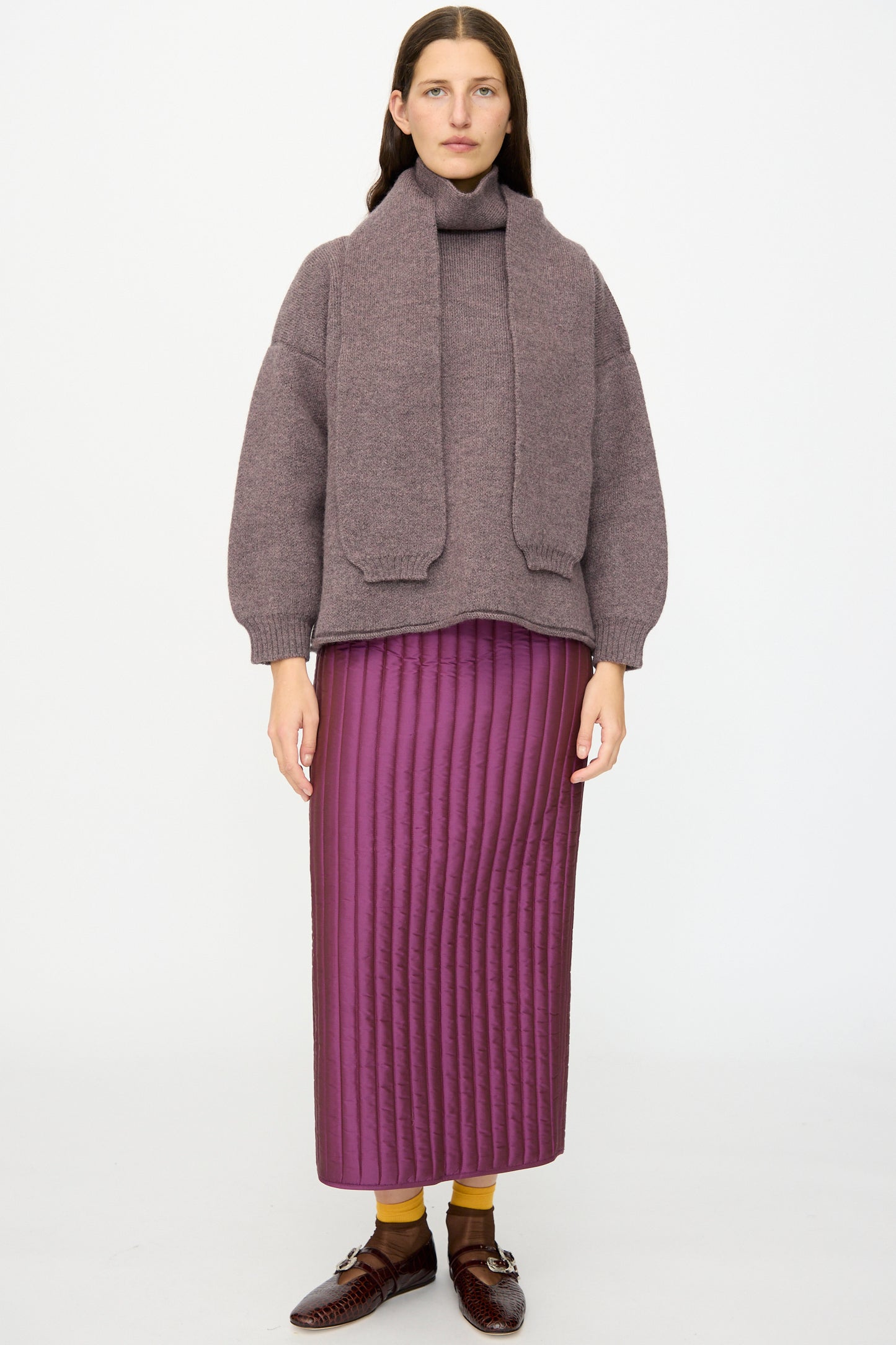 A person stands against a white background wearing the Cawley British Merino Wool Wrap Neck Pullover in Luna, paired with a matching scarf, a quilted purple skirt, yellow socks, and brown loafers.