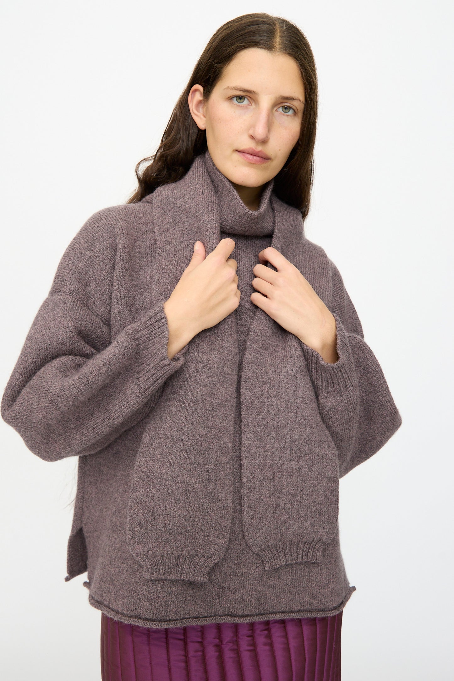 A woman with long brown hair wears the Cawley British Merino Wool Wrap Neck Pullover in Luna and a purple skirt, standing against a plain white background.