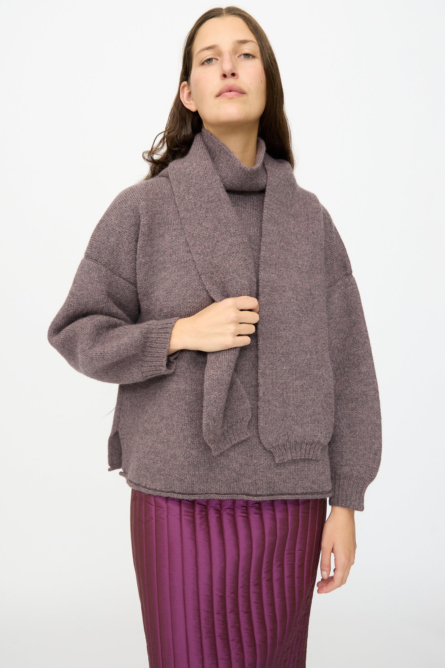 A woman stands against a plain background wearing the Cawley British Merino Wool Wrap Neck Pullover in Luna, paired with a matching scarf and a quilted purple skirt.