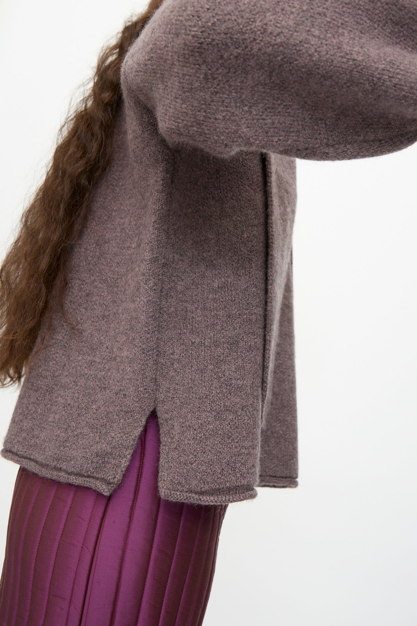 A person wears the Cawley British Merino Wool Wrap Neck Pullover in Luna, mauve, with side slits, over purple pleated pants, shown from mid-torso down against a plain background.