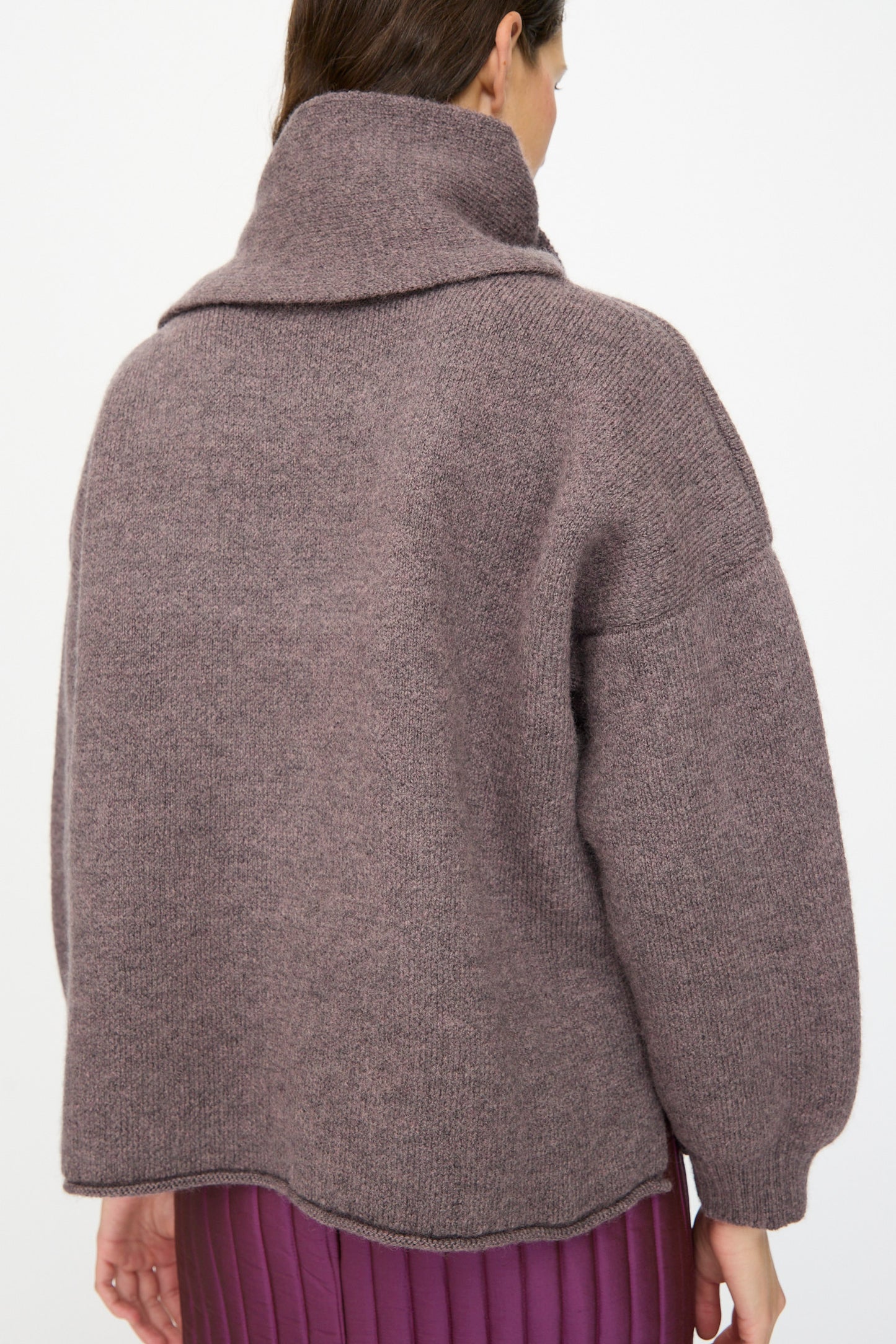 A woman is seen from the back wearing the Cawley British Merino Wool Wrap Neck Pullover in Luna, styled with a pleated purple skirt.