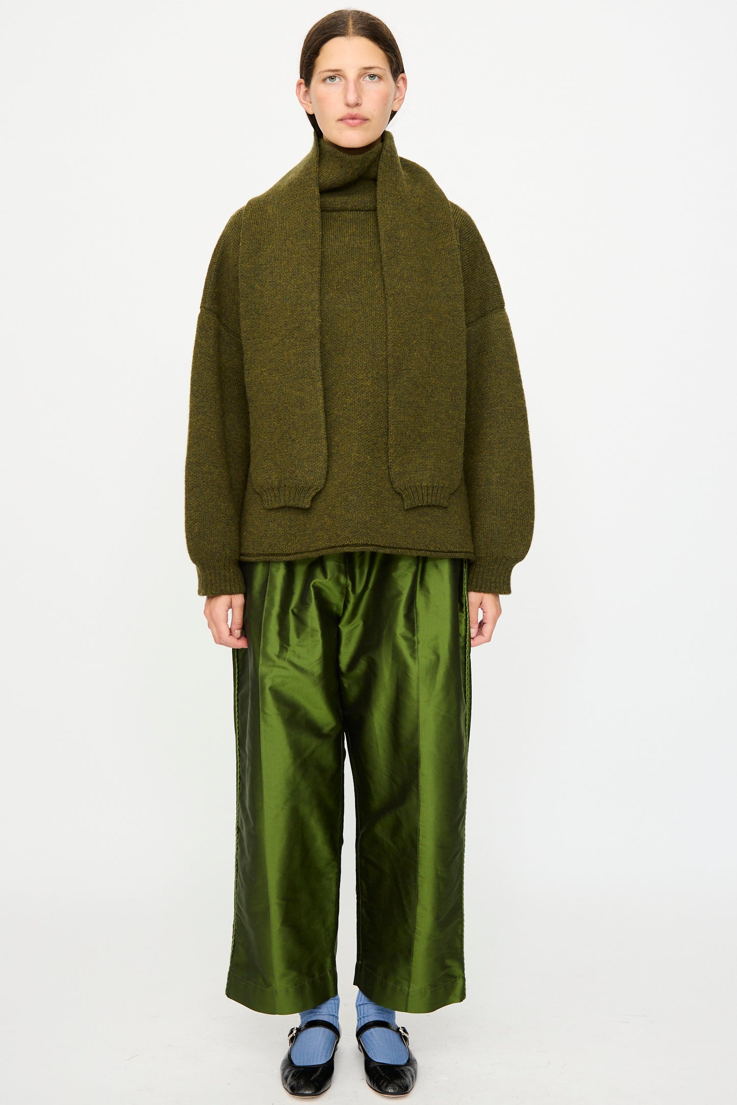 A person stands against a white background wearing the Cawley British Merino Wool Wrap Neck Pullover in Moss, made in London, styled with a matching scarf, shiny wide-leg green pants, black shoes, and blue socks.