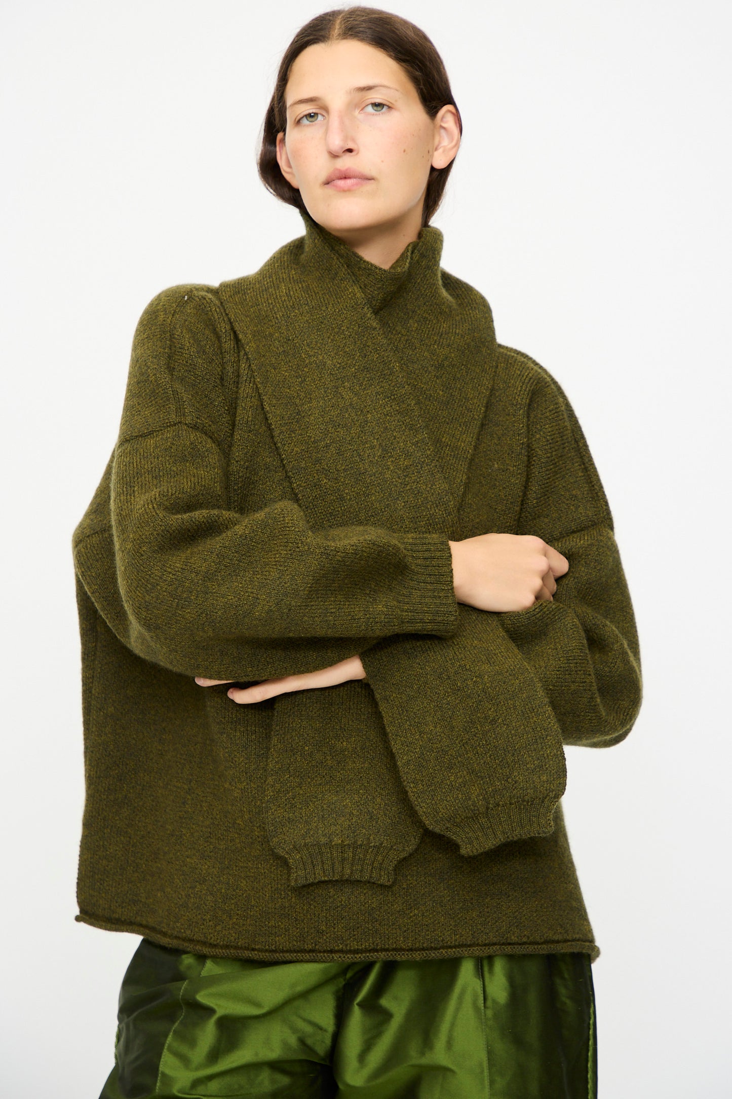 A person wears the Cawley British Merino Wool Wrap Neck Pullover in Moss, paired with a matching scarf and shiny green pants, standing against a plain white background.