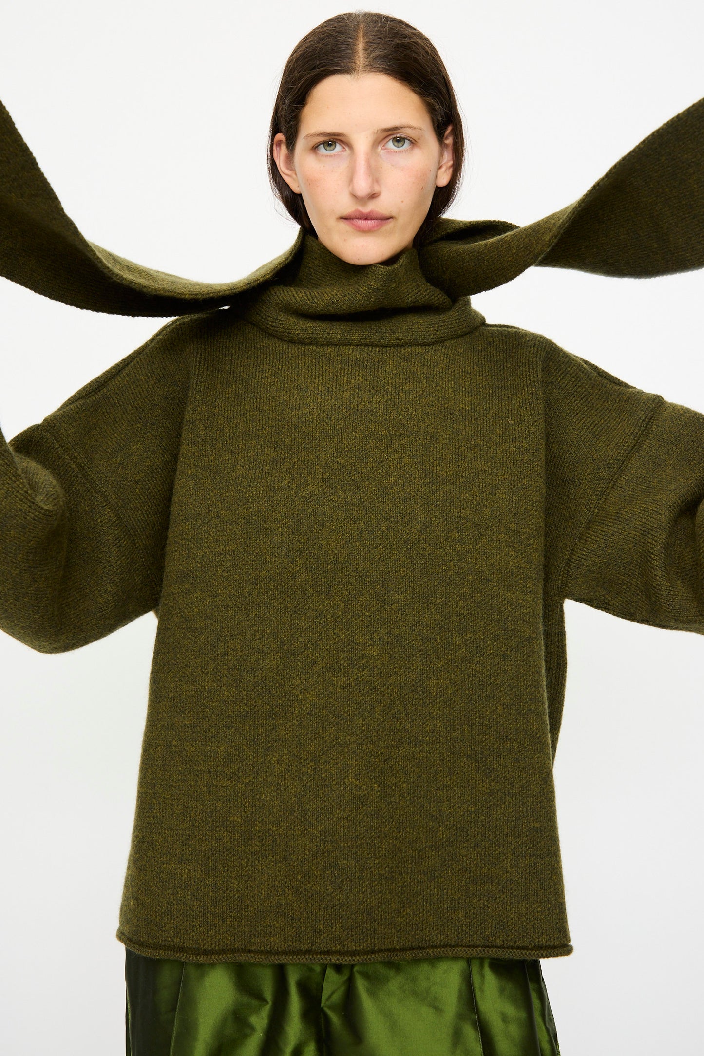 A person wears the Cawley British Merino Wool Wrap Neck Pullover in Moss, styled with a shiny green skirt, against a plain white background.