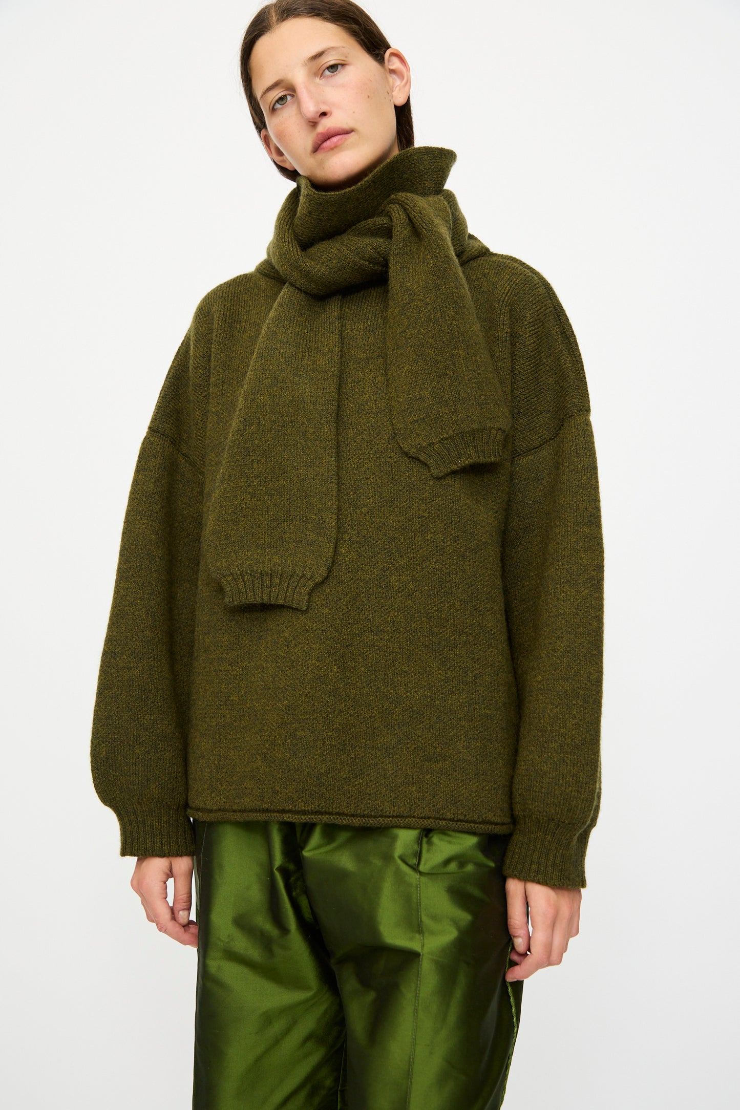 A person wearing the Cawley British Merino Wool Wrap Neck Pullover in Moss stands against a plain white background, paired with shiny green satin pants.