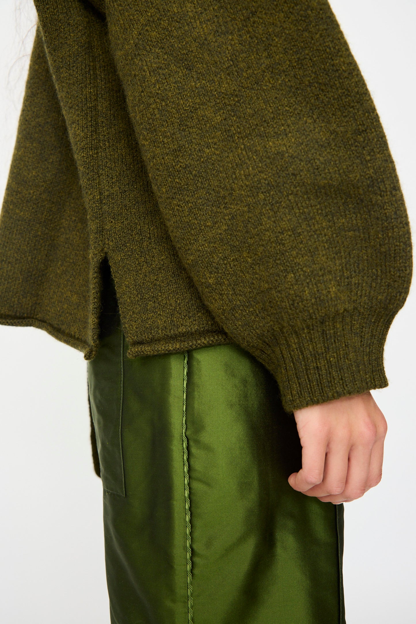 A person wearing the Cawley British Merino Wool Wrap Neck Pullover in Moss, a textured olive-green sweater with split hem, paired with a dark green satin skirt, shown from mid-torso to hand.