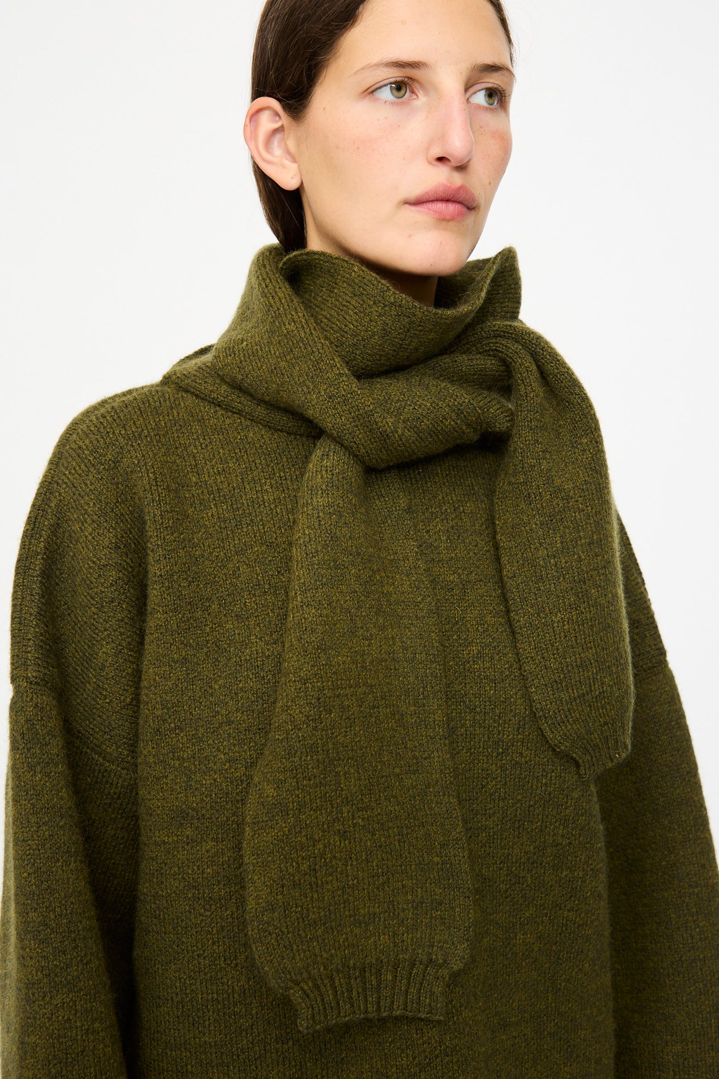 A person wears the Cawley British Merino Wool Wrap Neck Pullover in Moss, styled with the attached wrap neck, standing against a plain light background.