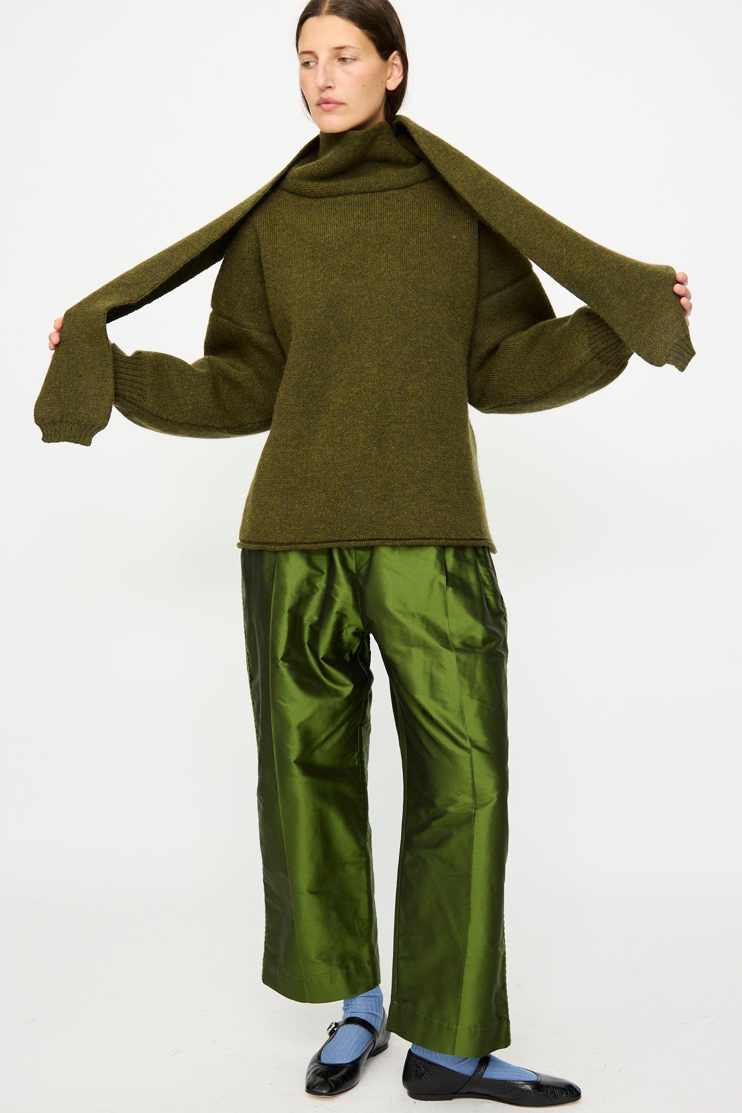 A person wearing the Cawley British Merino Wool Wrap Neck Pullover in Moss, styled with shiny green pants, blue socks, and black shoes, stands against a plain white background. Made in London.