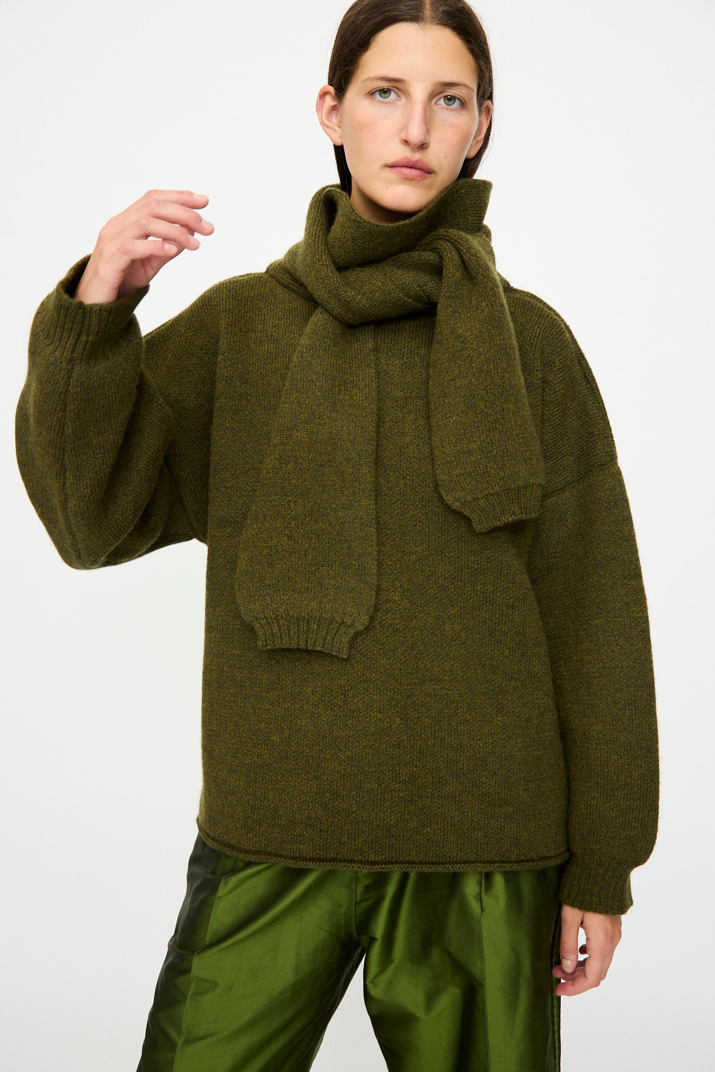 A person wears the Cawley British Merino Wool Wrap Neck Pullover in Moss, styled with a matching scarf and olive green pants, standing against a plain white background.