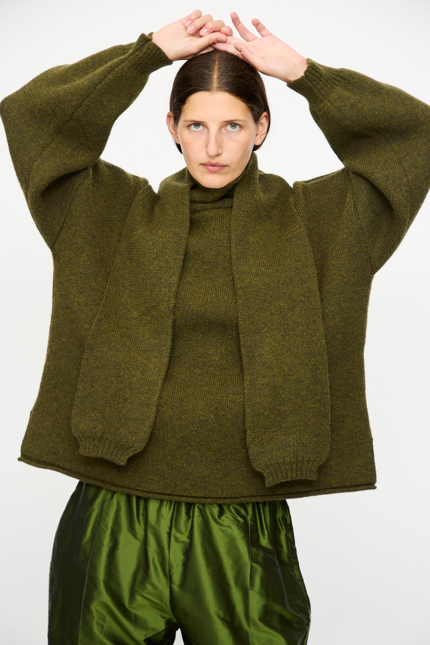 A person wears the Cawley British Merino Wool Wrap Neck Pullover in Moss with a scarf and shiny green pants, standing against a plain background with hands raised above their head.