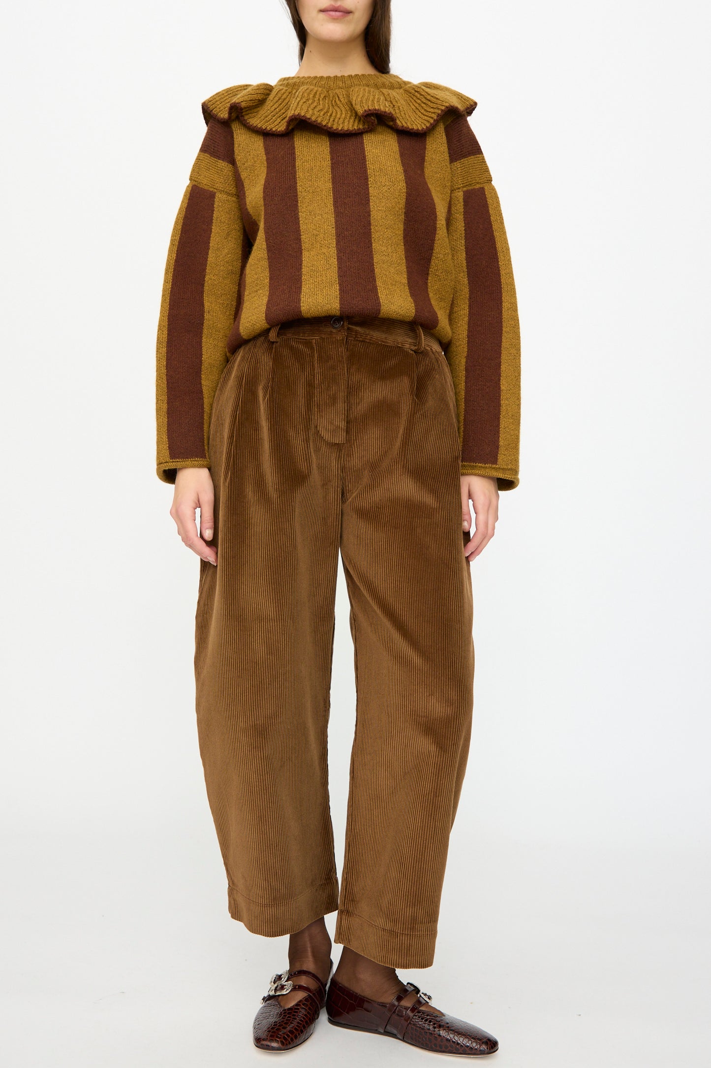 A person wearing Cawley's Cotton Wale Cord Robyn Trouser in Tan, paired with a mustard and brown striped ruffled-collar sweater and brown loafers, stands against a plain white background.