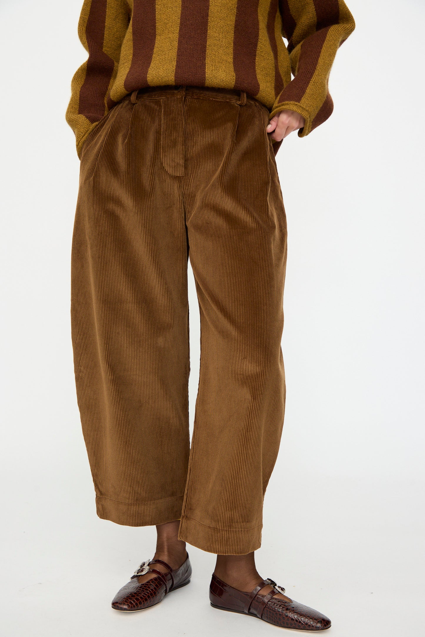 A person wears Cawley’s Cotton Wale Cord Robyn Trouser in Tan, paired with brown woven leather shoes and a brown and yellow striped sweater, standing against a plain white background.