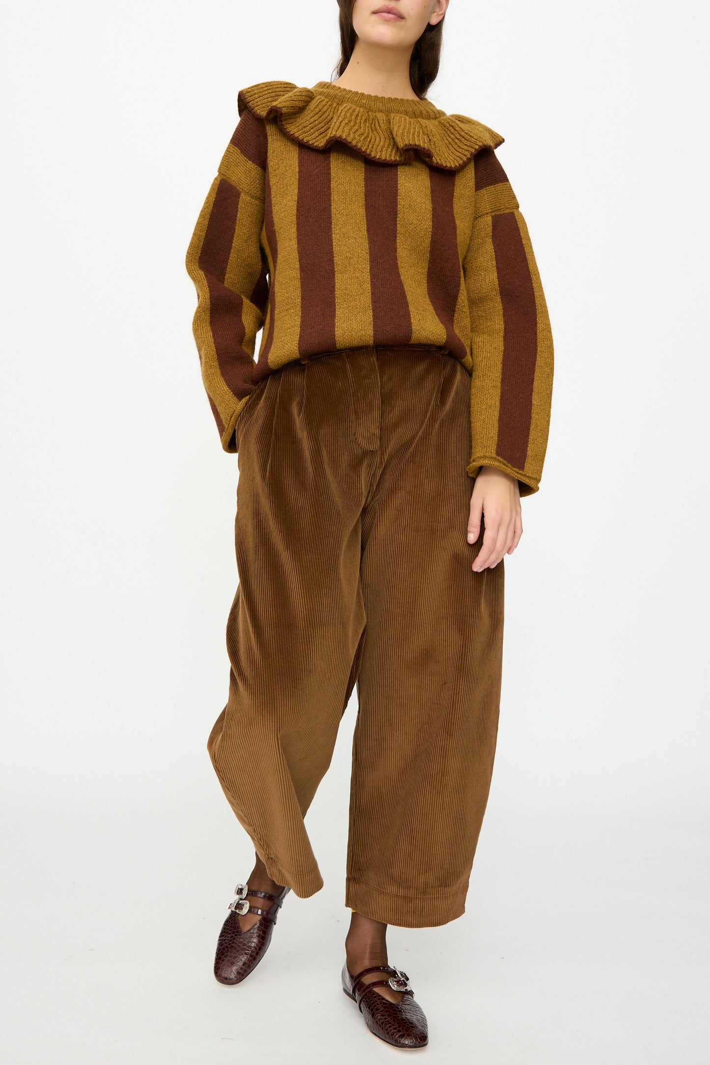 A person stands against a plain white background wearing Cawley’s Cotton Wale Cord Robyn Trouser in Tan, paired with a mustard and brown striped sweater, ruffled collar, and brown loafers.