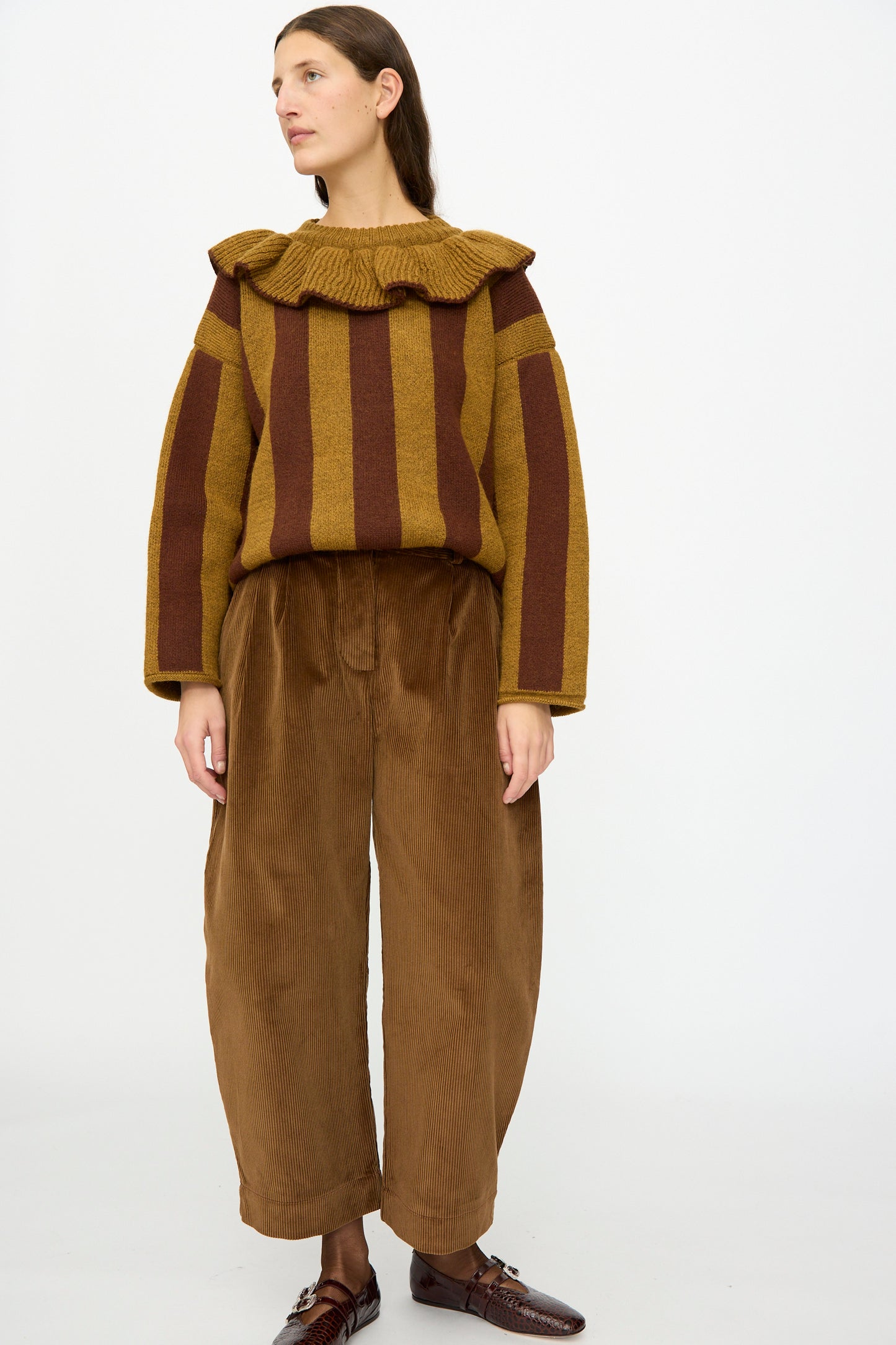 A person stands against a plain background wearing a brown and yellow striped sweater, brown shoes, and Cawley’s Cotton Wale Cord Robyn Trouser in Tan.