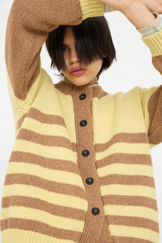 A person with a short dark bob haircut poses in a Cotton and Lambswool Textured Stripe Cardigan in Celery and Nutmeg from Cawley, one hand raised to their head.
