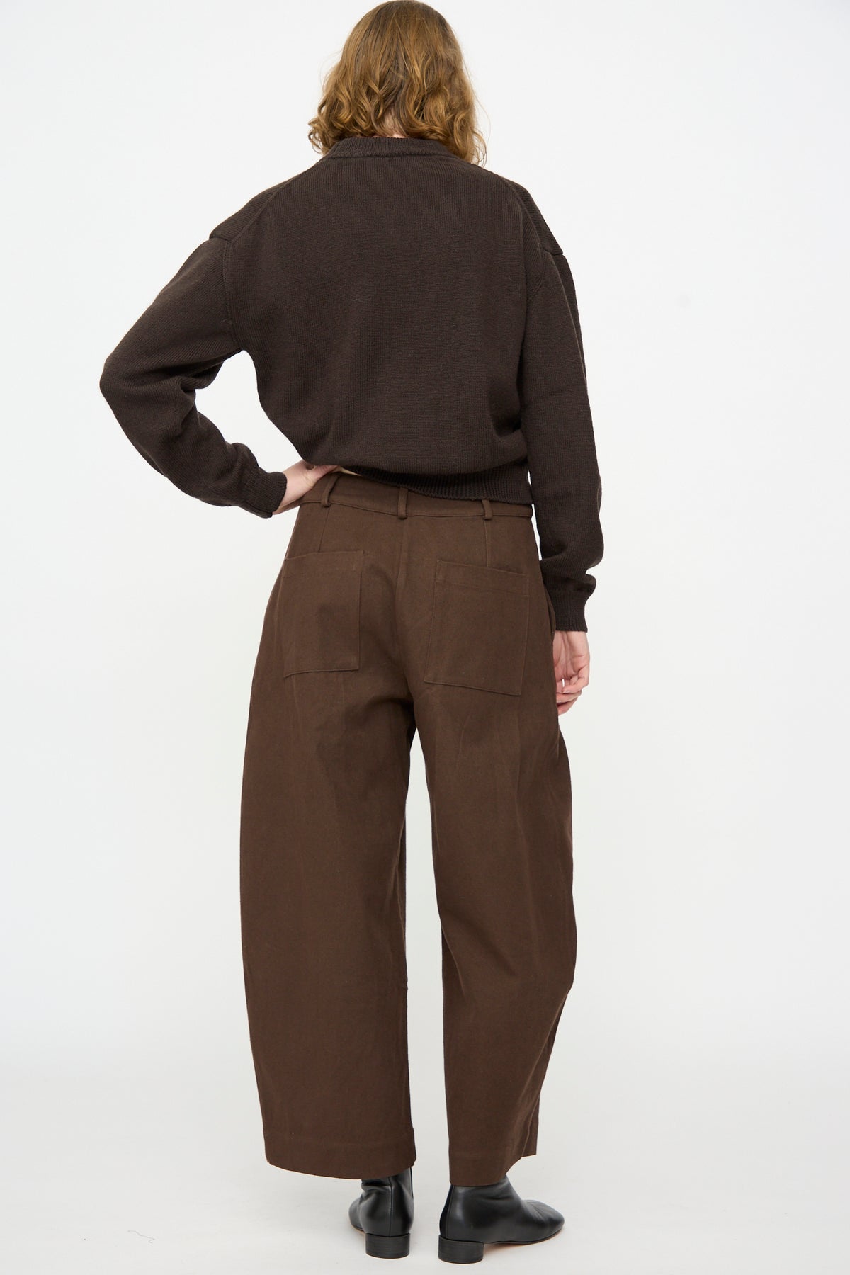 A person stands with their back to the camera against a plain white background, wearing a dark brown sweater, black shoes, and Cawley’s Japanese Gabardine Robyn Trouser in Espresso—featuring a relaxed fit and wide legs.