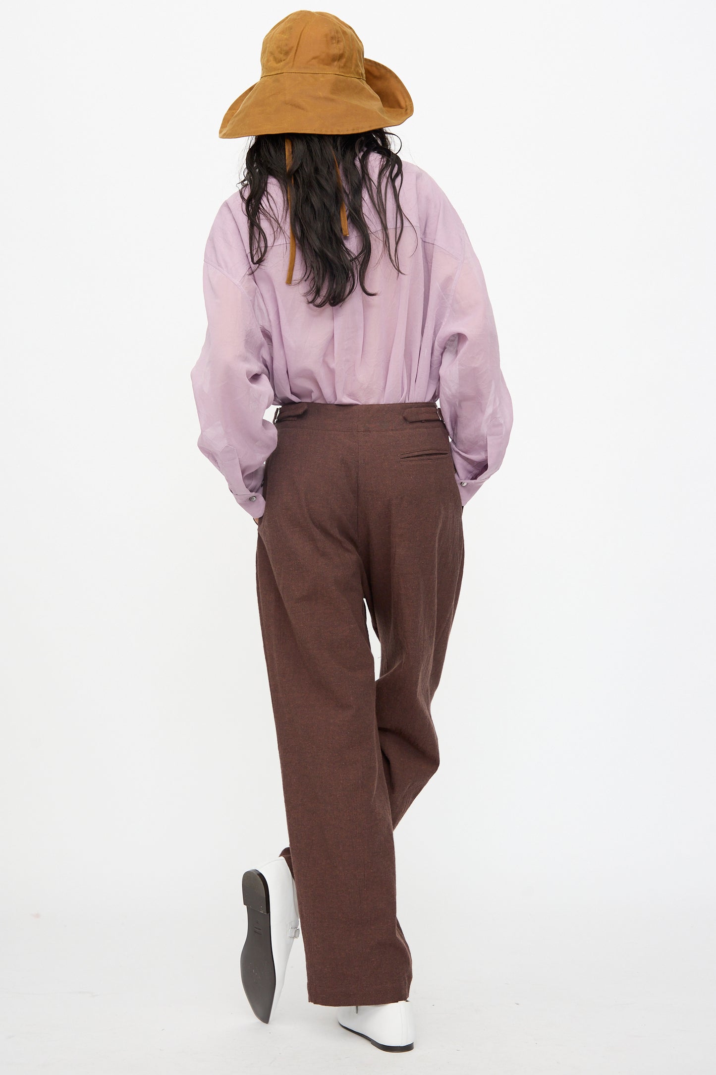Person facing away, wearing a brown wide-brim hat, loose lavender shirt, Cawley Japanese High Twisted Poplin Enya Trouser in Fog Purple with an adjustable waistband, and white shoes against a plain white background.