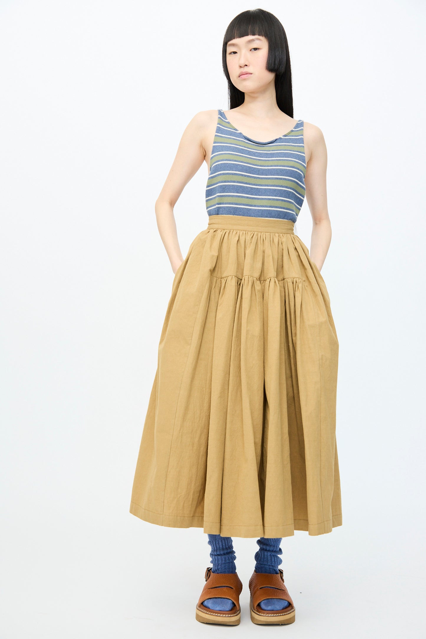 A person in a blue and gray striped sleeveless top, Cawley's Japanese Paper Cotton Panel Skirt in Nutmeg, blue socks, and brown sandals stands against a white background.