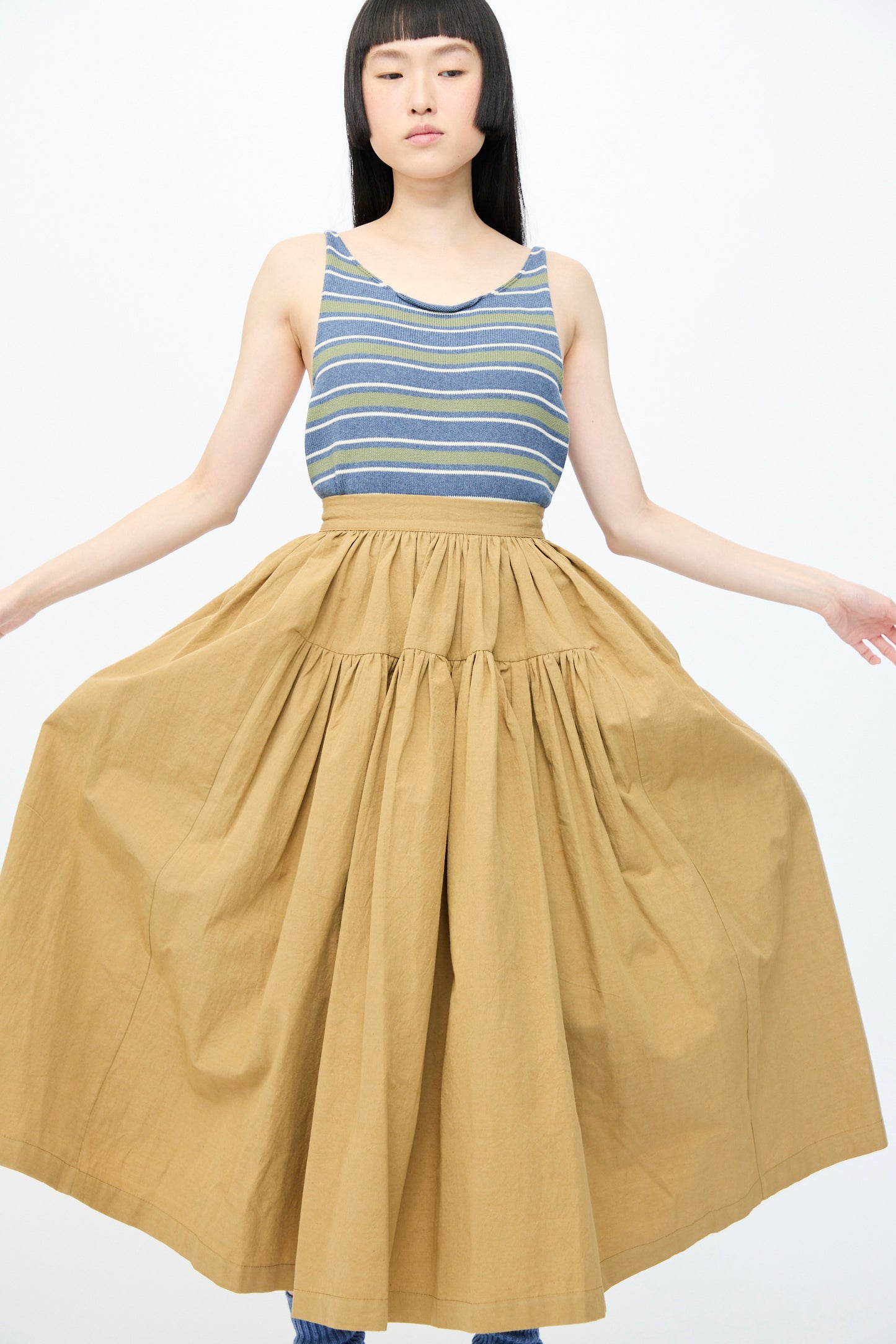 A person stands against a plain background wearing a striped tank top and the Cawley Japanese Paper Cotton Panel Skirt in Nutmeg, featuring a full, tan, gathered midi design with an A-line silhouette, their arms slightly extended to the sides.