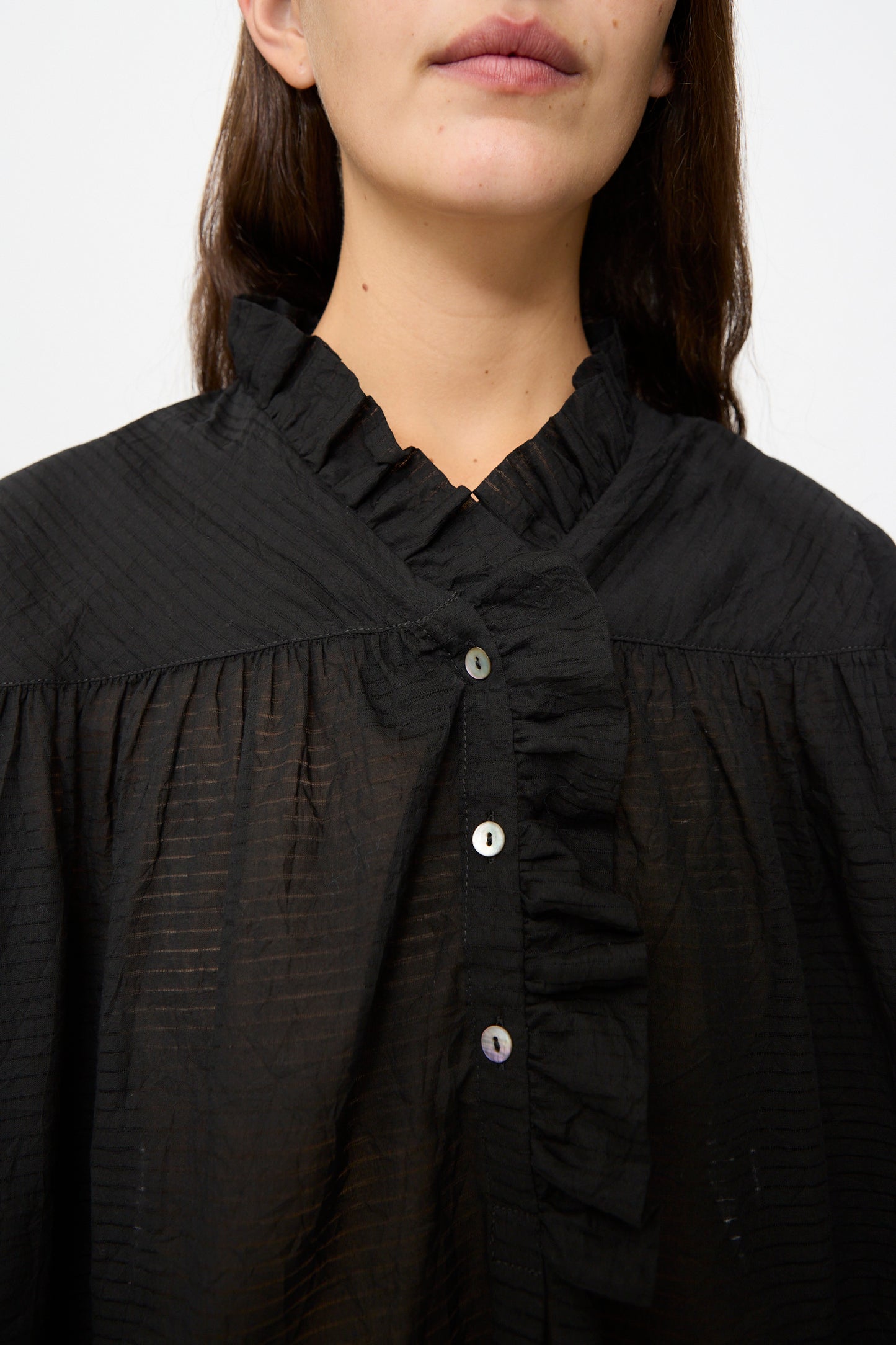 A person wearing the Cawley Japanese Sheer Stripe Cotton Sophie Shirt in Midnight, a black, textured long sleeve top with a ruffled collar and button details, is shown from shoulders to lips.