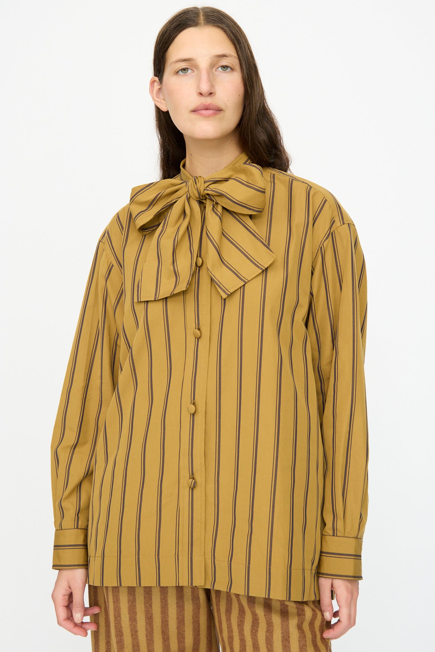 A person stands facing the camera wearing Cawley's Japanese Silk Cotton Hattie Shirt in Mustard and Brown Stripe, featuring long sleeves, a large collar bow, and matching striped pants.