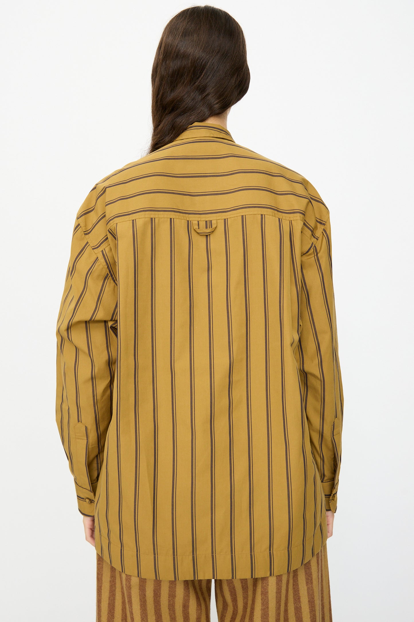 A person wears the Cawley Japanese Silk Cotton Hattie Shirt in Mustard and Brown Stripe, seen from the back with brown patterned pants. This long sleeve shirt is crafted in London.