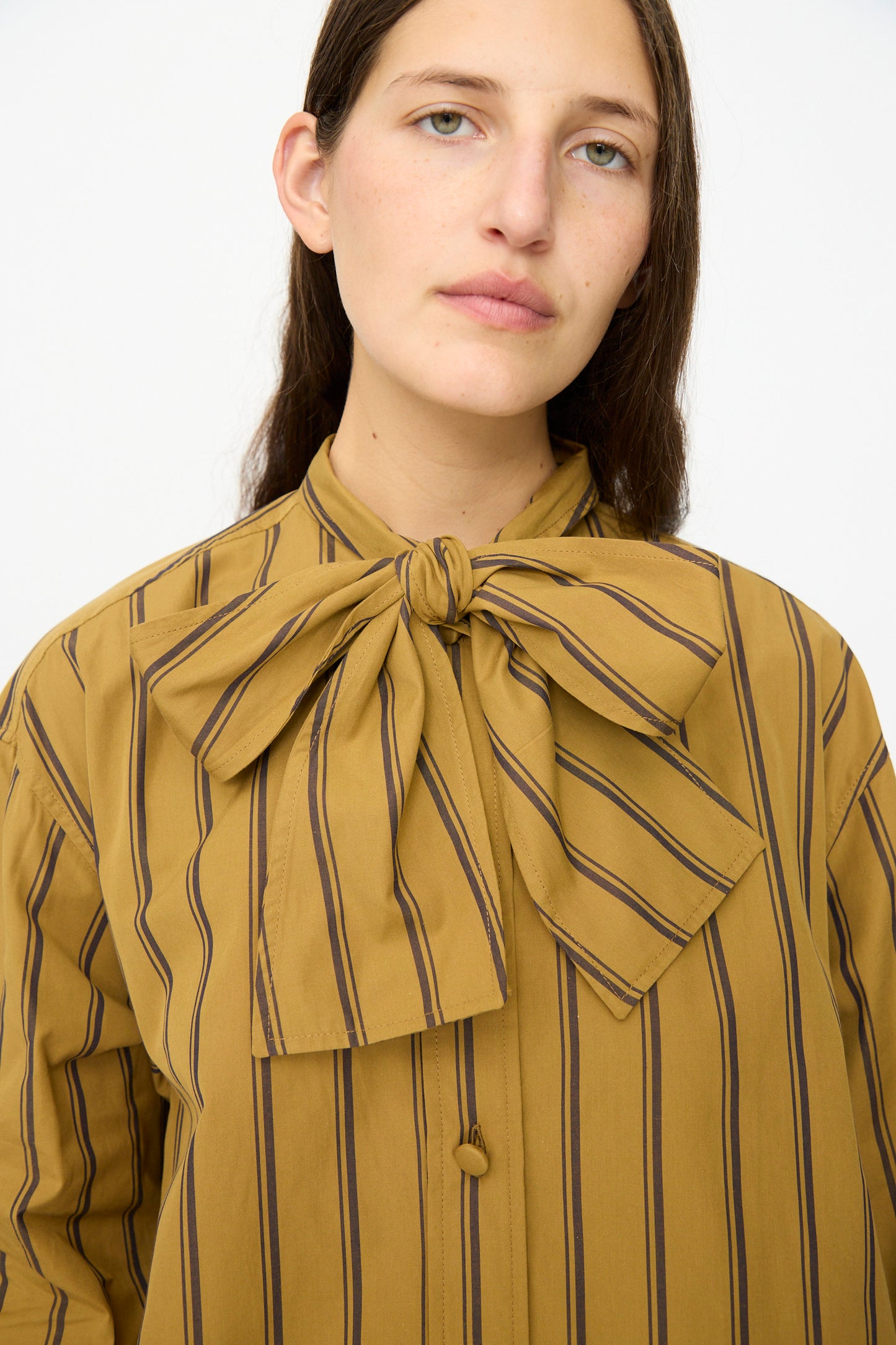 A woman stands against a plain white background wearing the Cawley Japanese Silk Cotton Hattie Shirt in mustard and brown stripe, featuring a large collar bow.