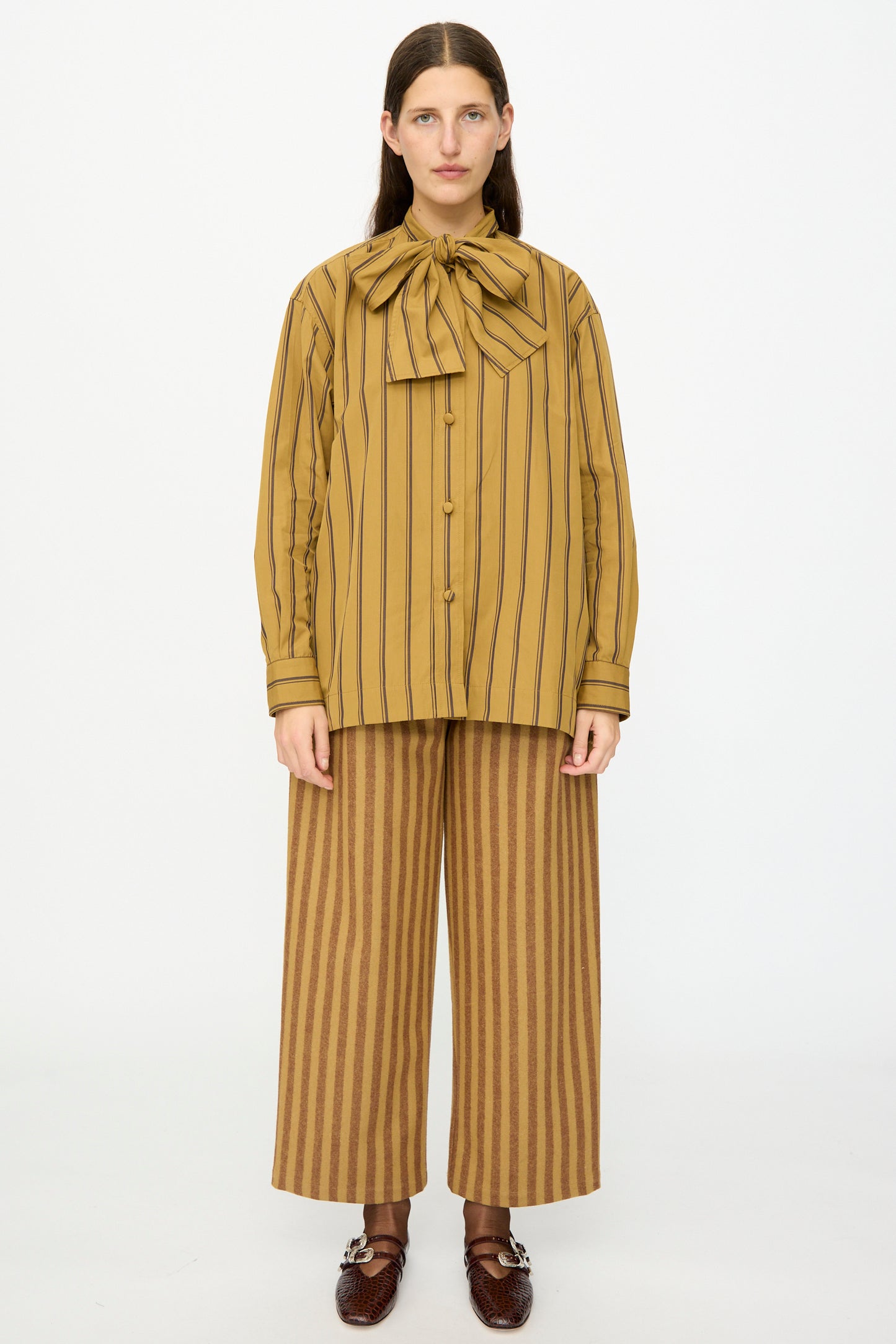 A person stands facing forward, wearing the Cawley Japanese Silk Cotton Hattie Shirt in Mustard and Brown Stripe, paired with matching wide-leg pants and brown woven shoes.