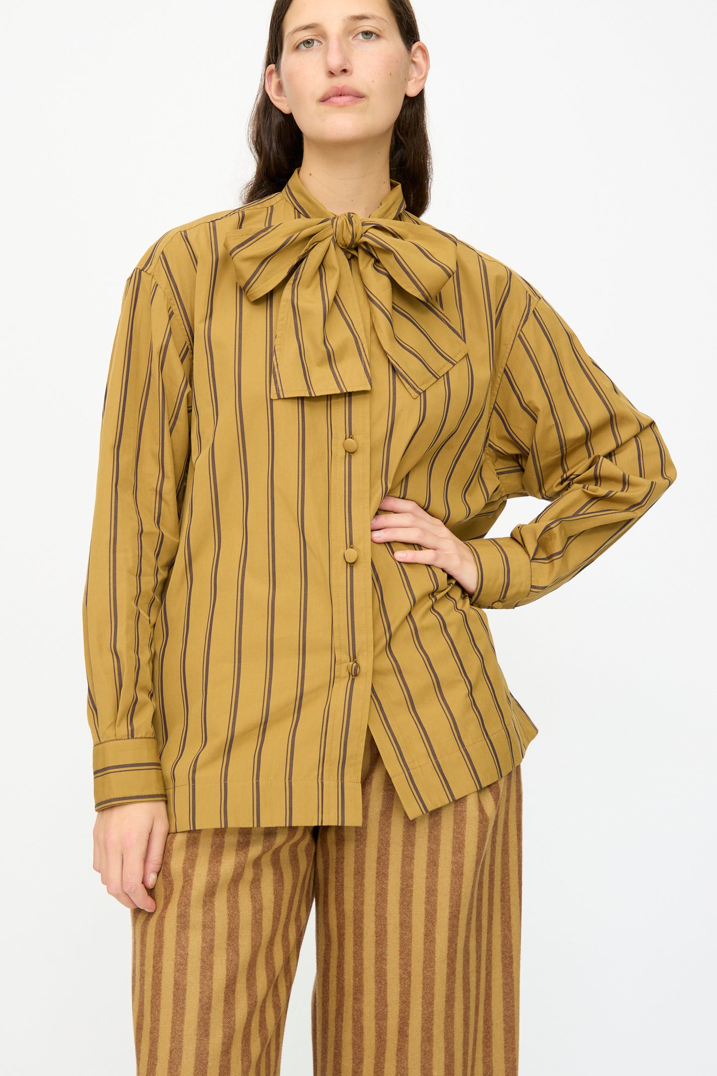 A person wears the Cawley Japanese Silk Cotton Hattie Shirt in mustard and brown stripe, featuring long sleeves and a large neck bow, paired with striped pants, against a plain background. Made in London.