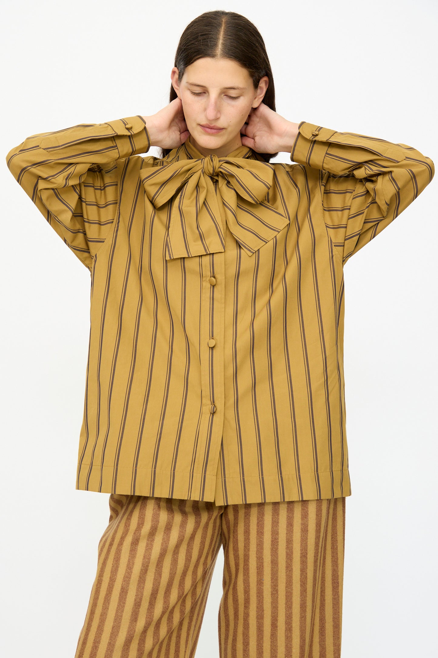 A person wearing Cawley’s Japanese Silk Cotton Hattie Shirt in Mustard and Brown Stripe, featuring vertical stripes and a large neck bow, stands with hands behind their neck. This long-sleeve shirt is made in London.