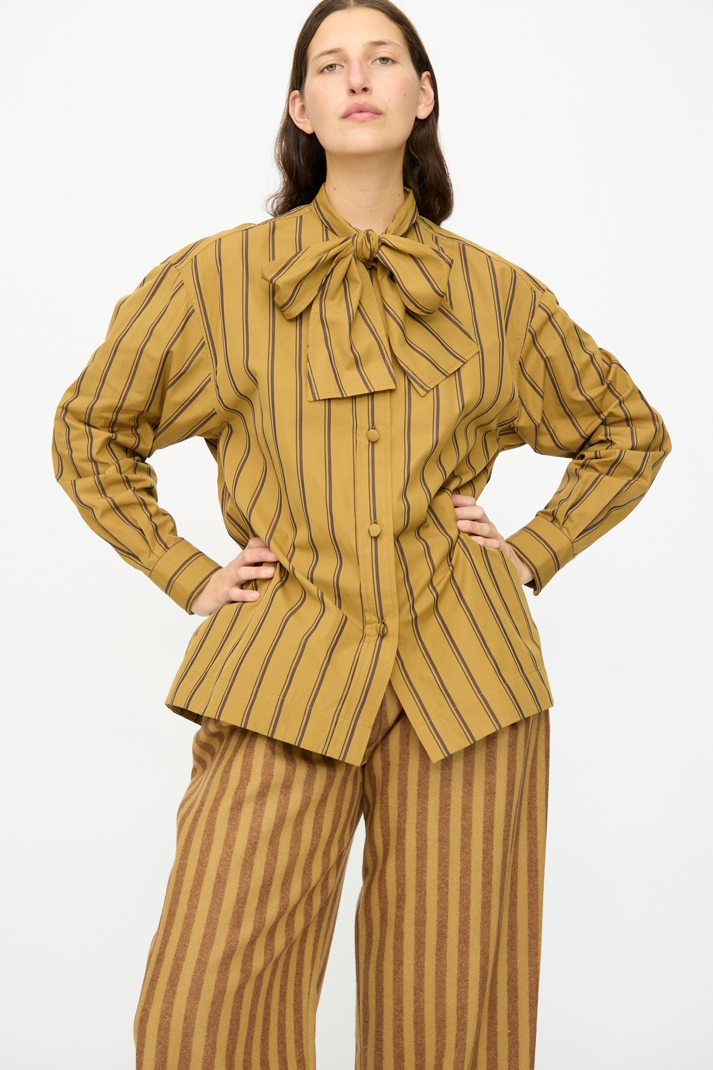 A person wears the Cawley Japanese Silk Cotton Hattie Shirt in Mustard and Brown Stripe, featuring black and brown vertical stripes, a large neck bow, and long sleeves, paired with wide-leg pants. Made in London.