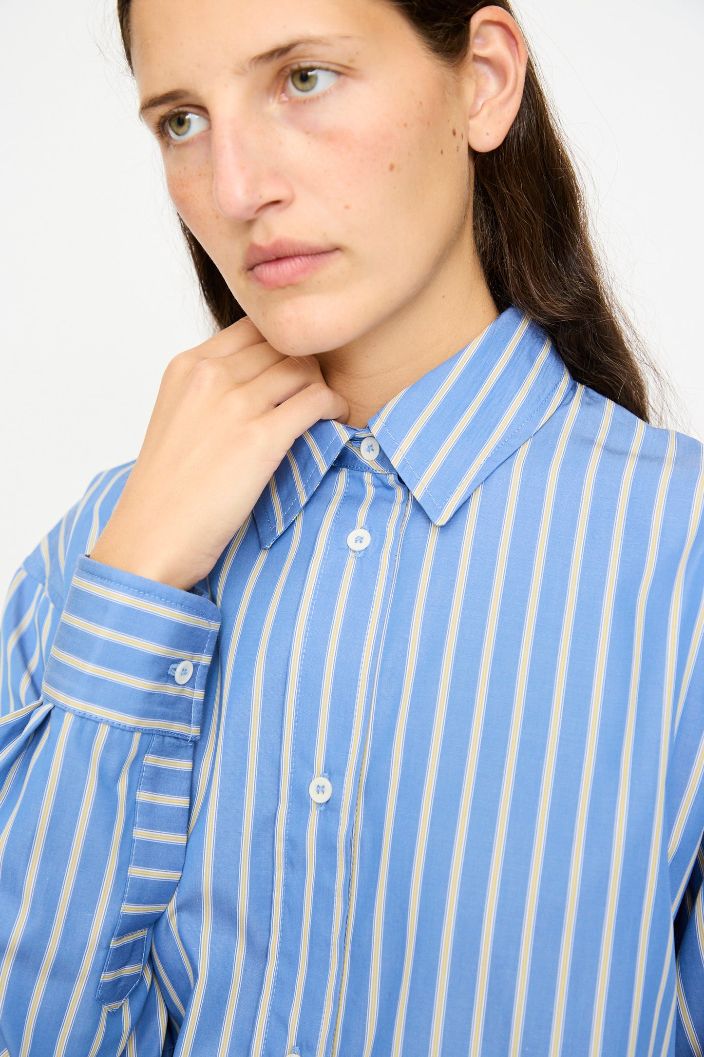 A person with long brown hair wears the Cawley Japanese Striped Dennis Shirt in Blue Dandelion Stripe, looking slightly to the side and touching their collar.