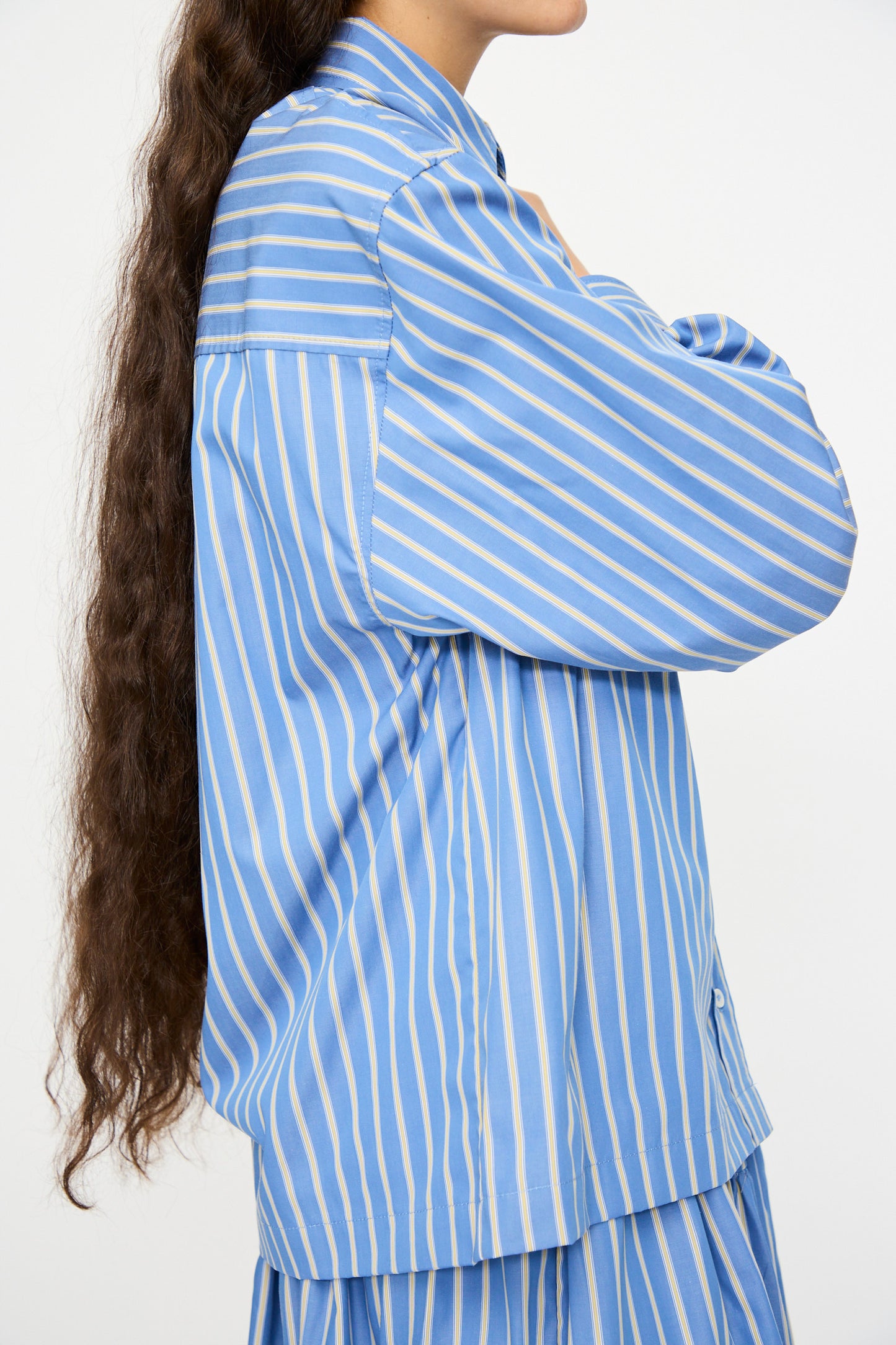 A person with long, wavy brown hair wears the Cawley Japanese Striped Dennis Shirt in Blue Dandelion Stripe, paired with a matching skirt, facing sideways against a plain background.