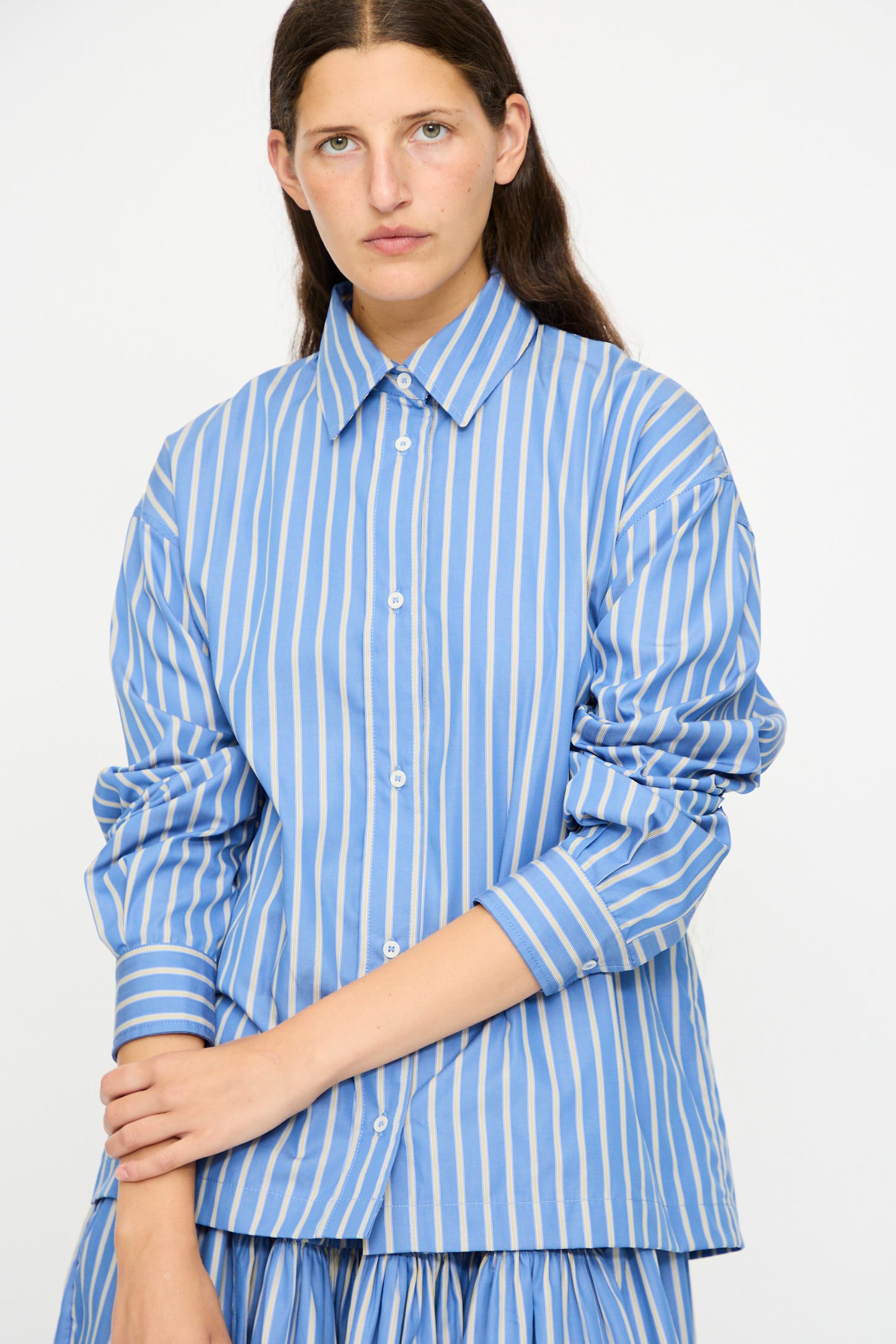 A woman wears the Cawley Japanese Striped Dennis Shirt in Blue Dandelion Stripe, a loose-fit cotton shirt with long sleeves and blue and white vertical stripes, standing against a plain light background.