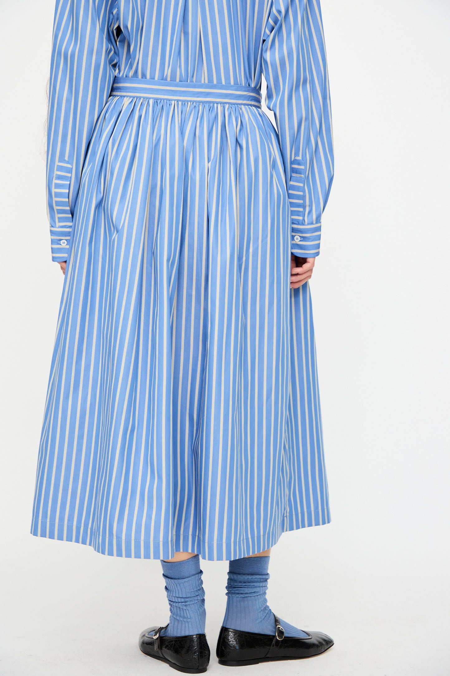 A person wears the Cawley Japanese Striped Shirting Panel Skirt in Dandelion, styled with matching socks and black flats. The A-line silhouette is photographed from the back against a plain background. Made in London.