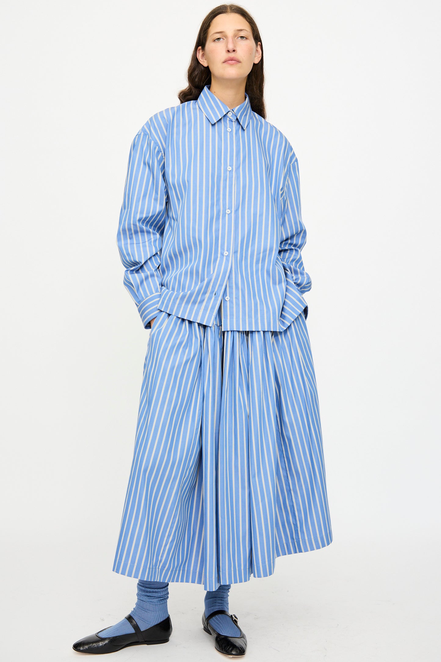 A person stands against a plain white background wearing the Cawley Japanese Striped Shirting Panel Skirt in Dandelion, paired with a matching button-up shirt, blue socks, and black shoes.