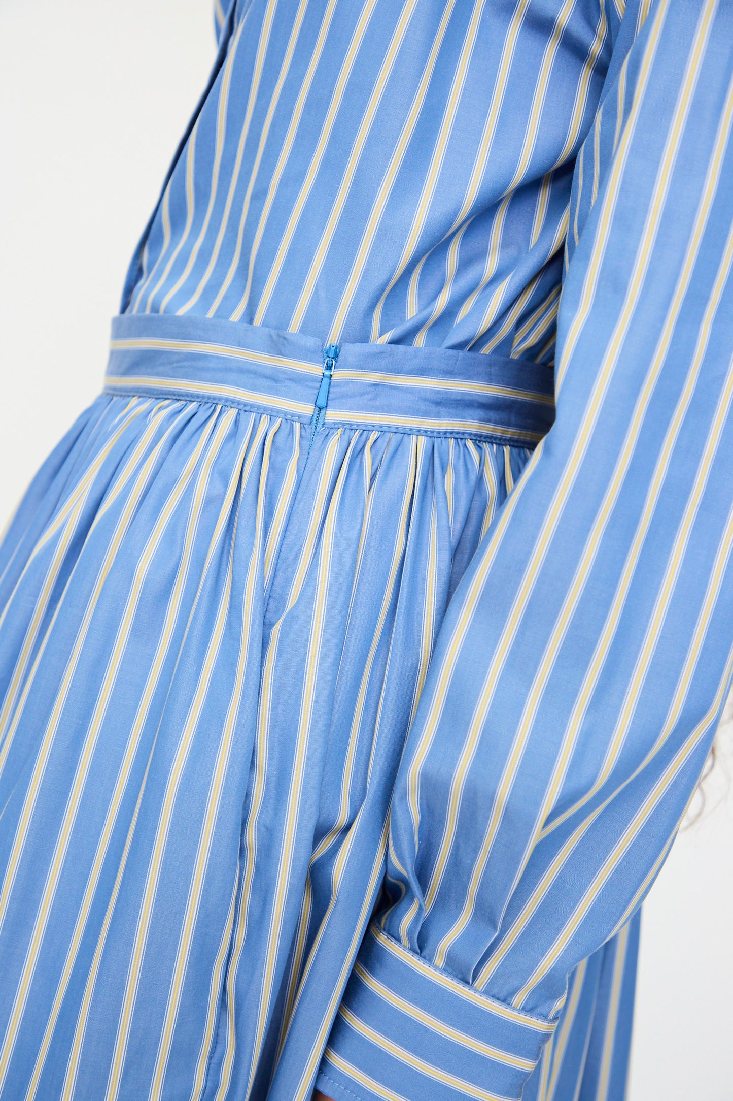Close-up of a person wearing Cawley's Japanese Striped Shirting Panel Skirt in Dandelion, featuring blue and white stripes, a belted waist, long sleeves, and an A-line silhouette.