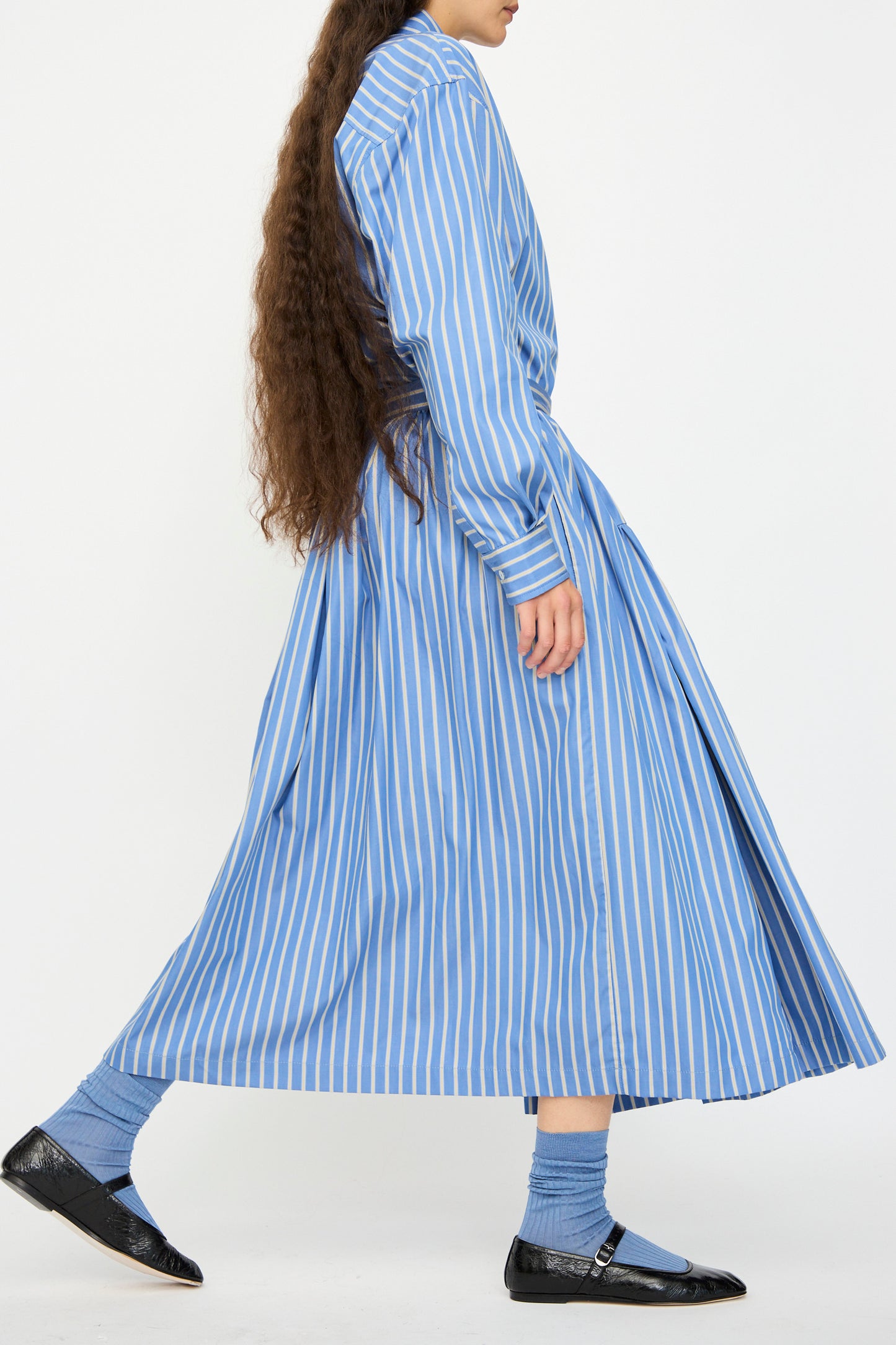 A person with long brown hair wears Cawley's Japanese Striped Shirting Panel Skirt in Dandelion, blue socks, and black Mary Jane shoes while walking against a white background.