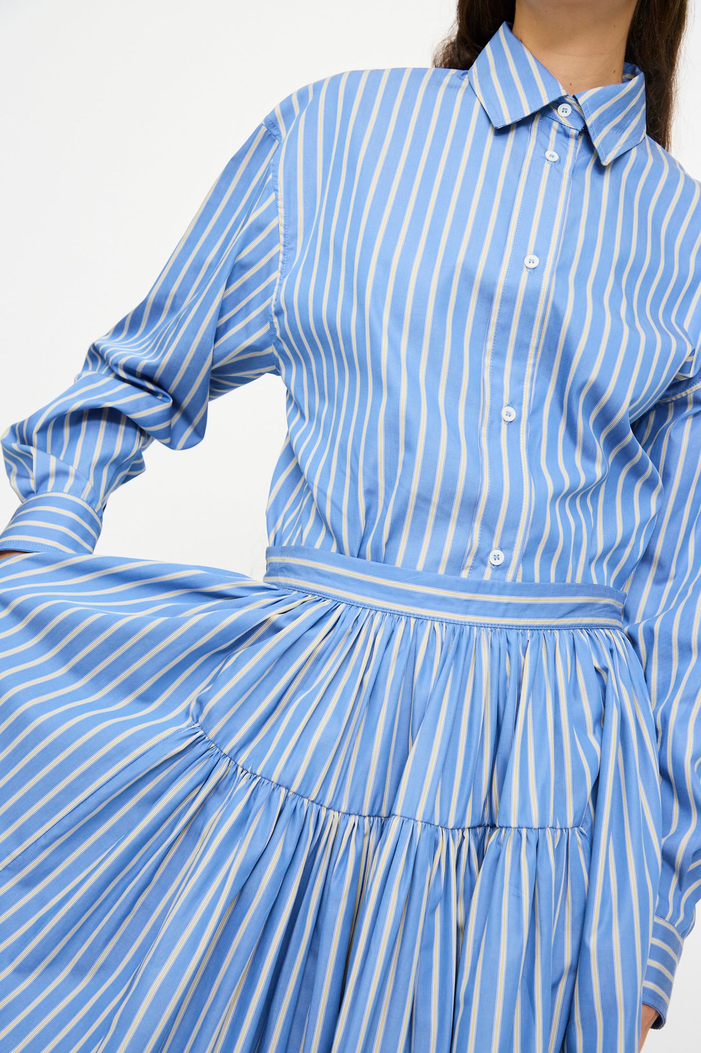 A person wears Cawley's Japanese Striped Shirting Panel Skirt in Dandelion, a chic A-line piece made in London, paired with a blue and white vertically striped button-up shirt, shown from shoulders to knees.