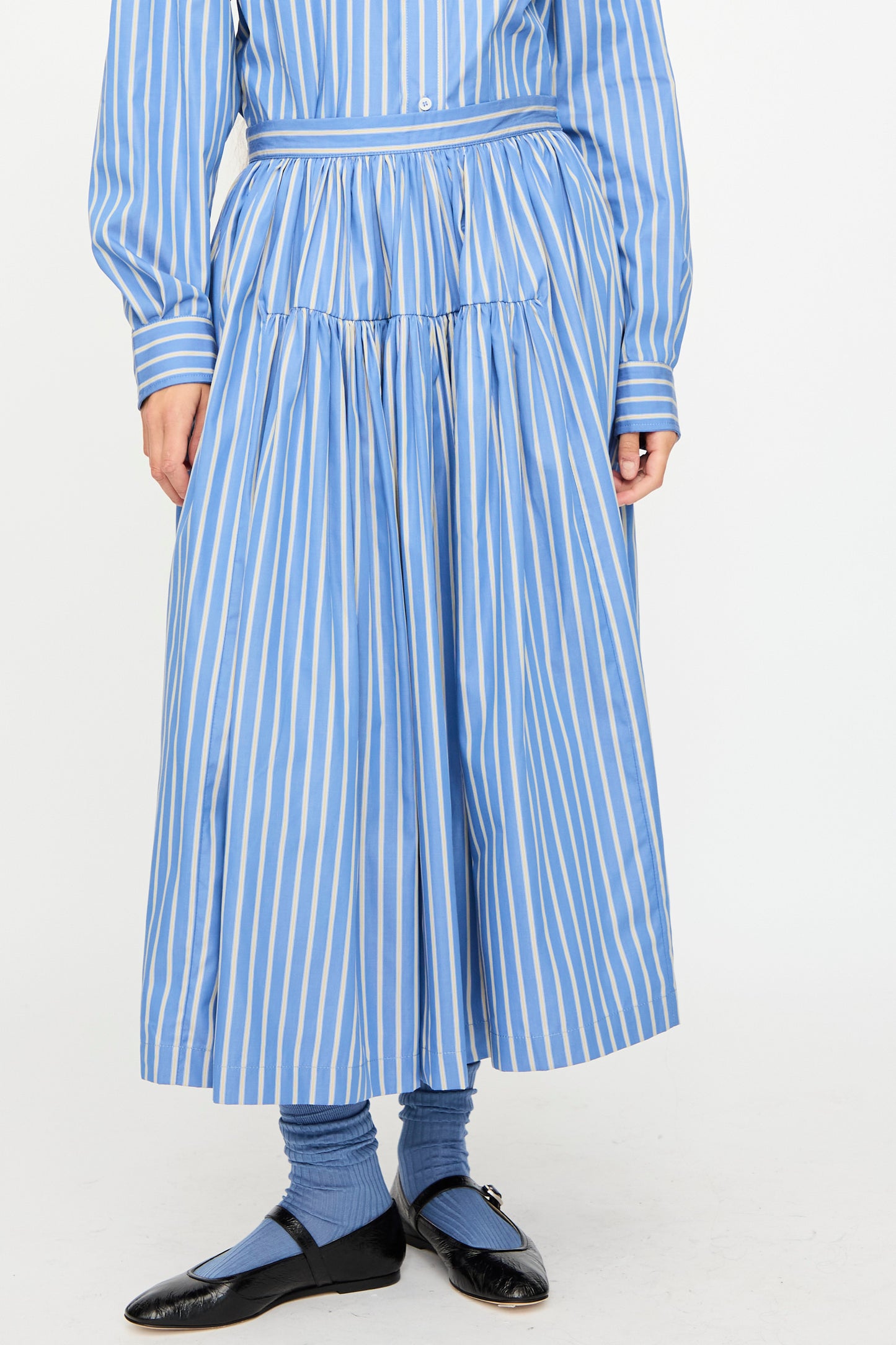 Person wearing a Cawley Japanese Striped Shirting Panel Skirt in Dandelion with a matching shirt, blue socks, and black shoes stands against a plain background.
