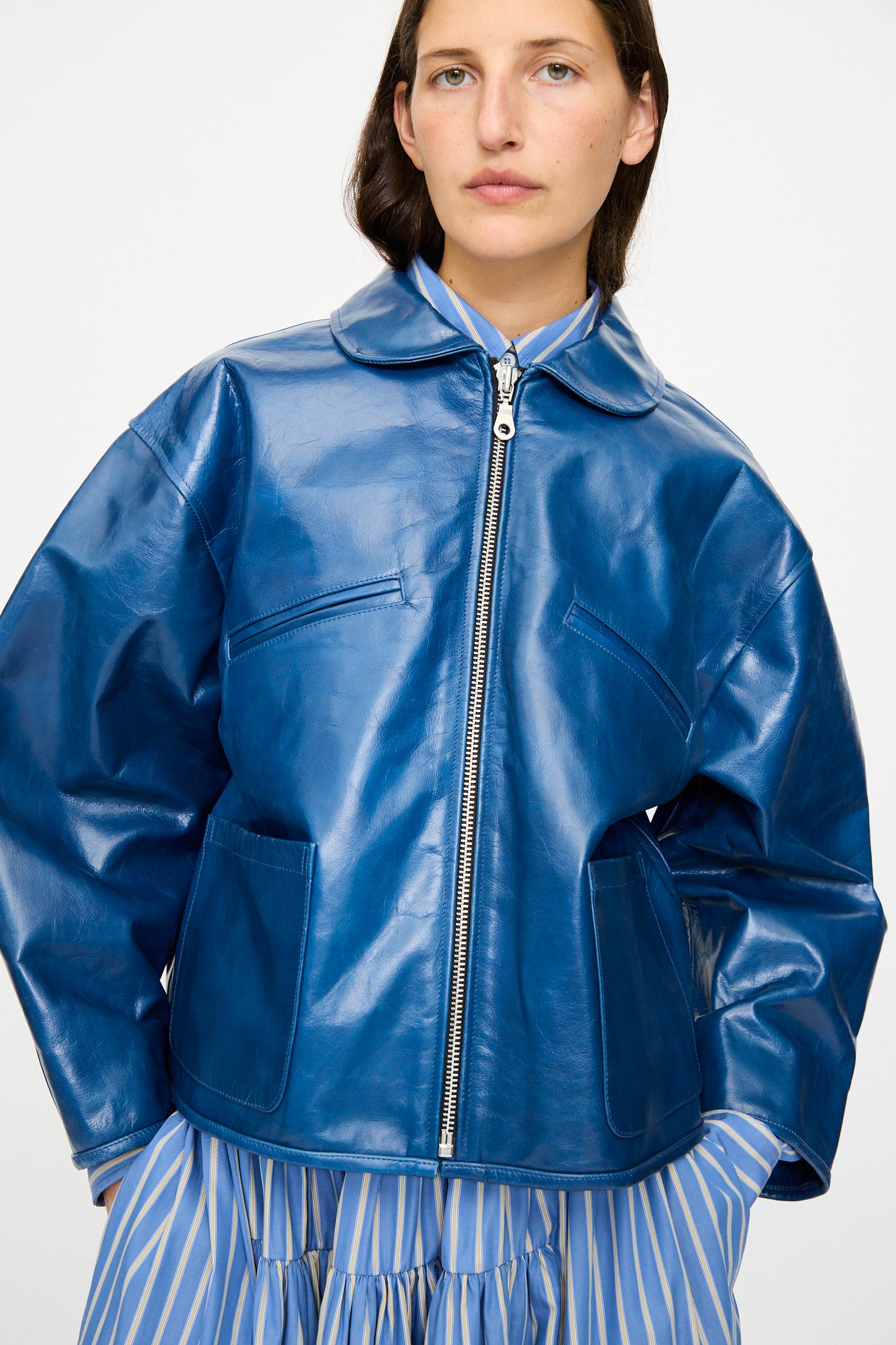 A person wears the Cawley Leather Flying Jacket in Canada Blue, styled in an oversized fit over a blue and white striped dress, standing against a plain background.