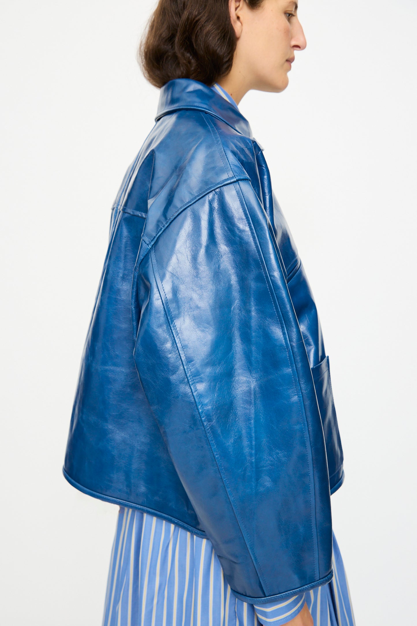 A person in profile wears the Cawley Leather Flying Jacket in Canada Blue—an oversized, shiny blue aviator style—over a blue and white striped shirt, set against a plain light background.