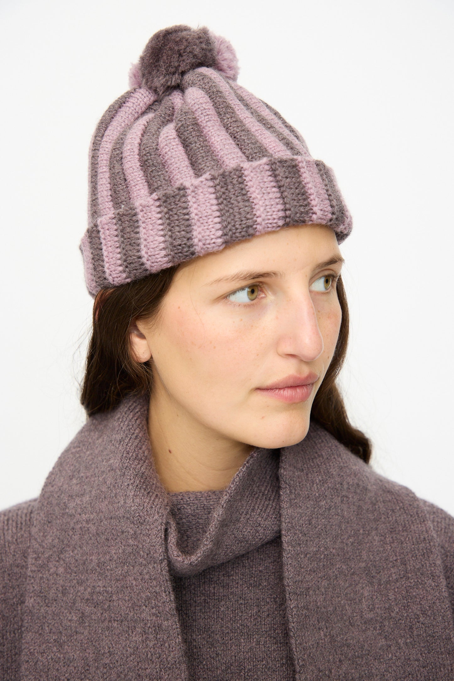 A person wears the Cawley Merino Wool Pleated Bobble Hat in Brezo and Luna, paired with a matching purple scarf and sweater, gazing to the side against a plain background.