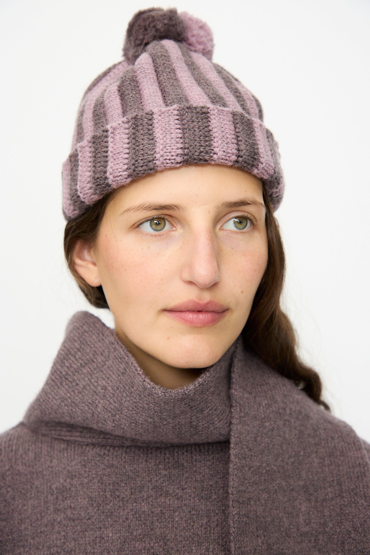 A person with light skin and long brown hair wears the Cawley Merino Wool Pleated Bobble Hat in Brezo and Luna, looking slightly to the right.