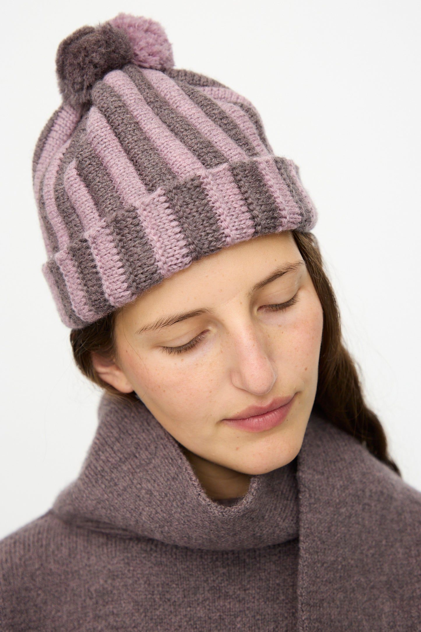 A woman in a Cawley Merino Wool Pleated Bobble Hat in Brezo and Luna gazes downward, her cozy knit accessories standing out against a plain background.