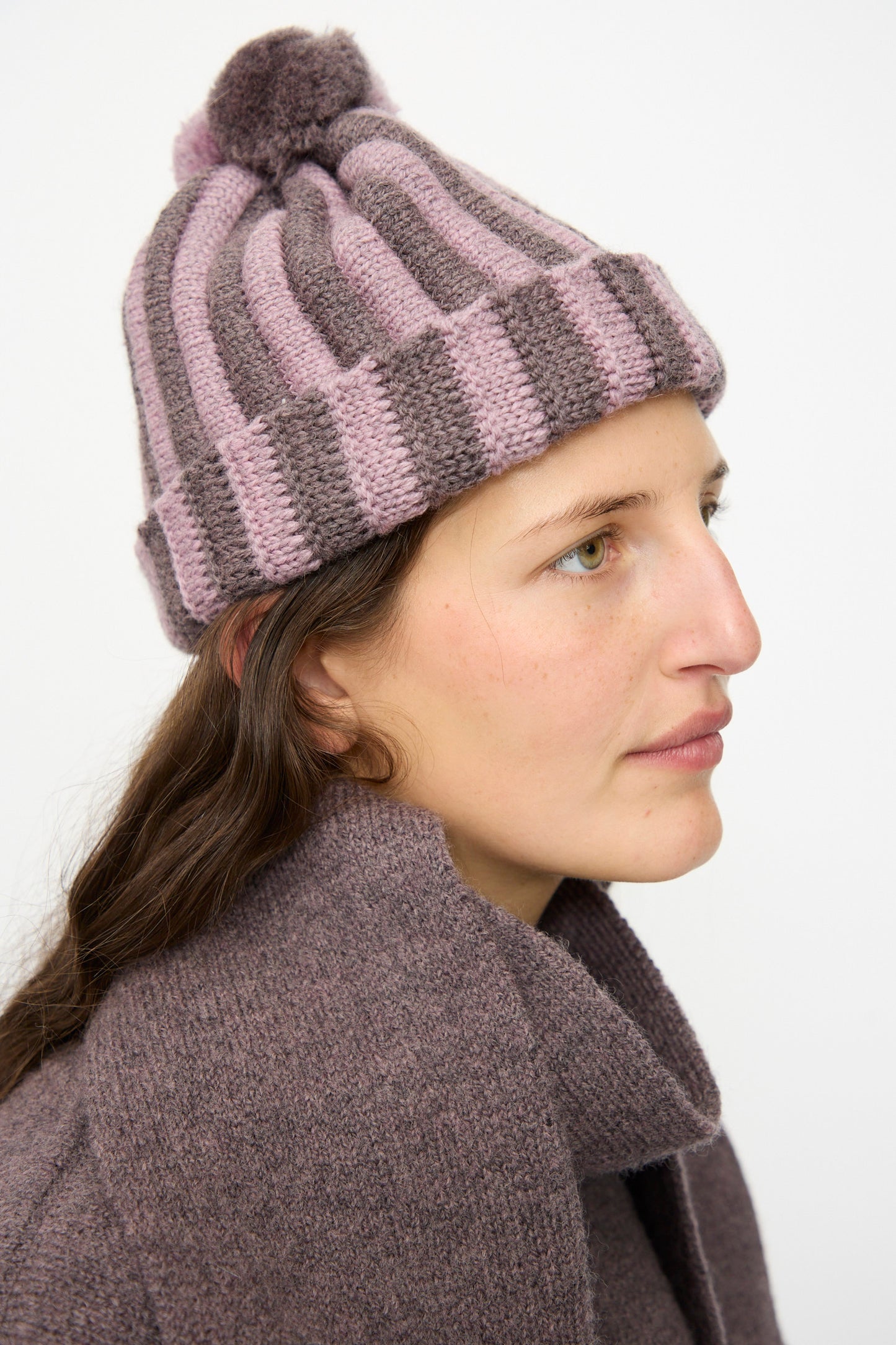 A woman in a Cawley Merino Wool Pleated Bobble Hat in Brezo and Luna, featuring a pom-pom, looks to the side in matching outerwear against a plain background.