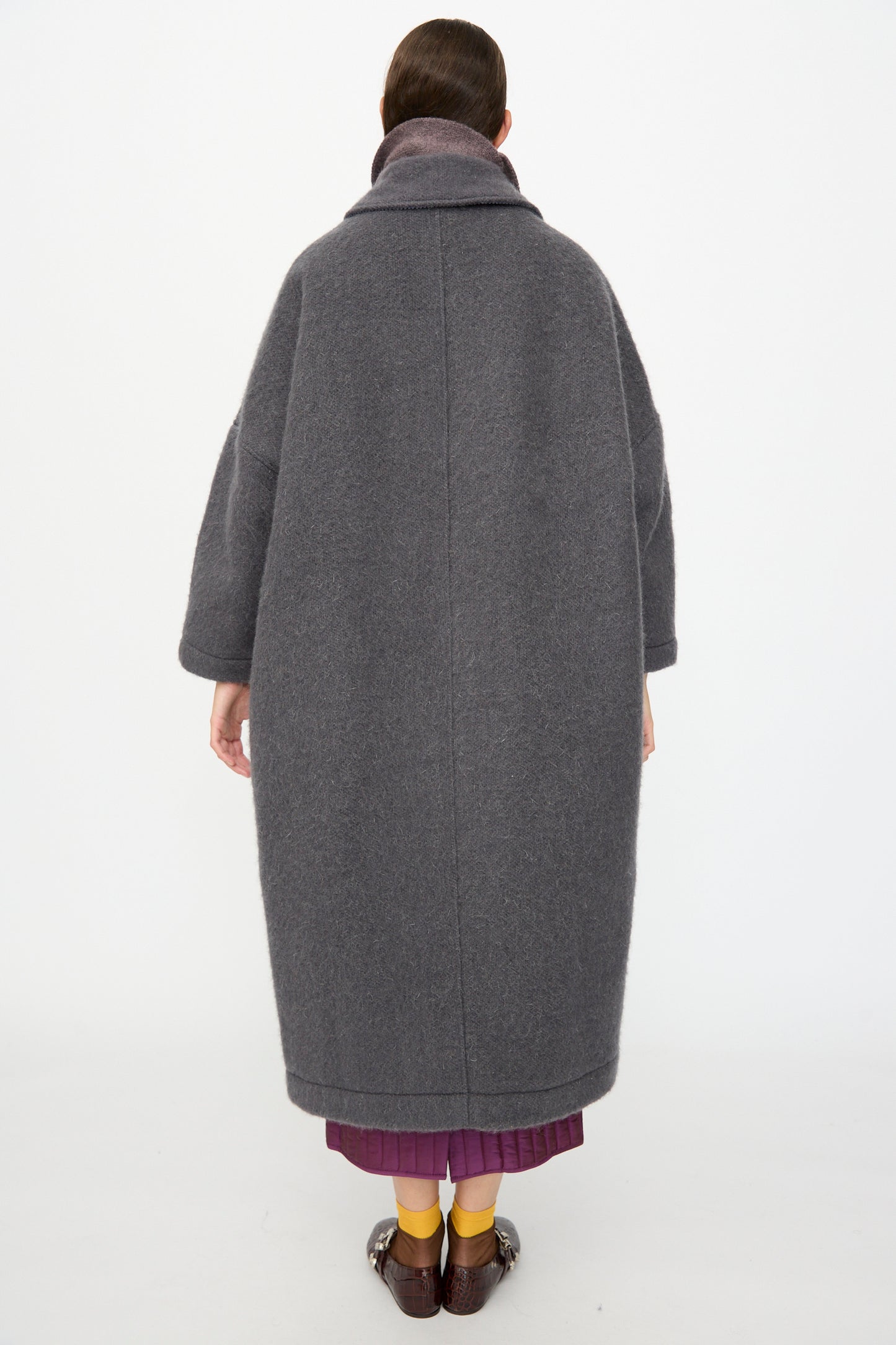 A person stands facing away, wearing the Cawley Portuguese Hairy Wool Faro Coat in Slate, paired with a purple skirt, yellow socks, and brown shoes, against a plain white background.