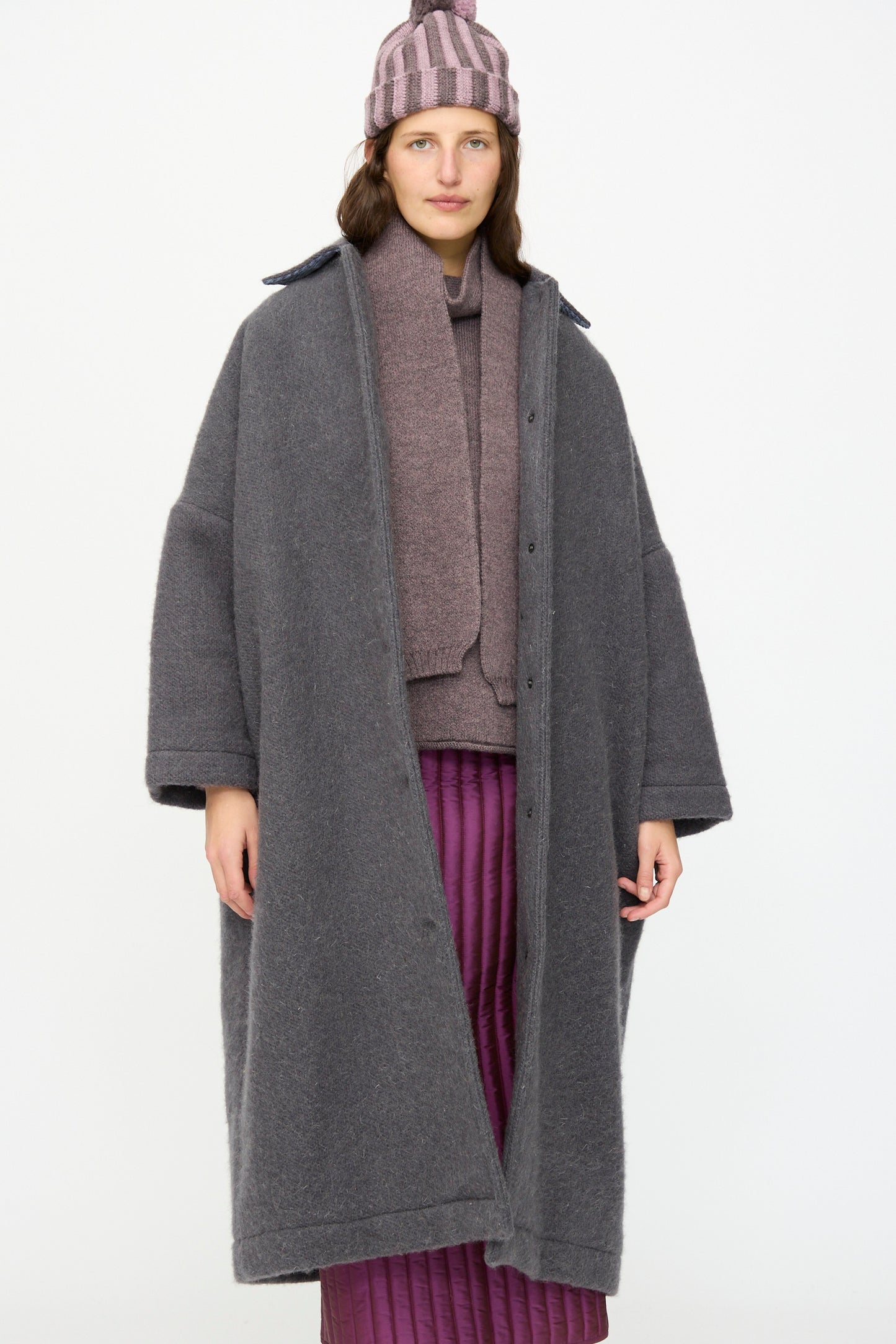 A person wears the Cawley Portuguese Hairy Wool Faro Coat in Slate, paired with a mauve sweater, scarf, striped pink beanie, and a quilted purple skirt, standing against a plain white background.