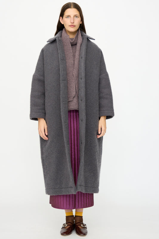 A person stands against a plain background wearing Cawley's Portuguese Hairy Wool Faro Coat in Slate, a mauve scarf, purple pleated skirt, yellow socks, and brown shoes.